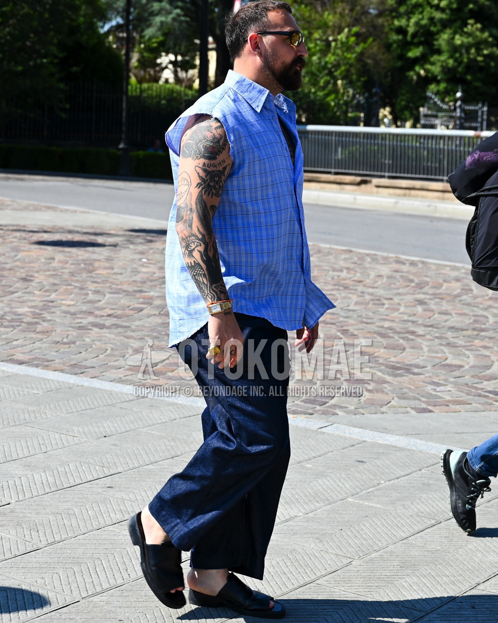 Men's spring summer outfit with clear plain sunglasses, light blue check shirt, navy plain slacks, black shower sandals.