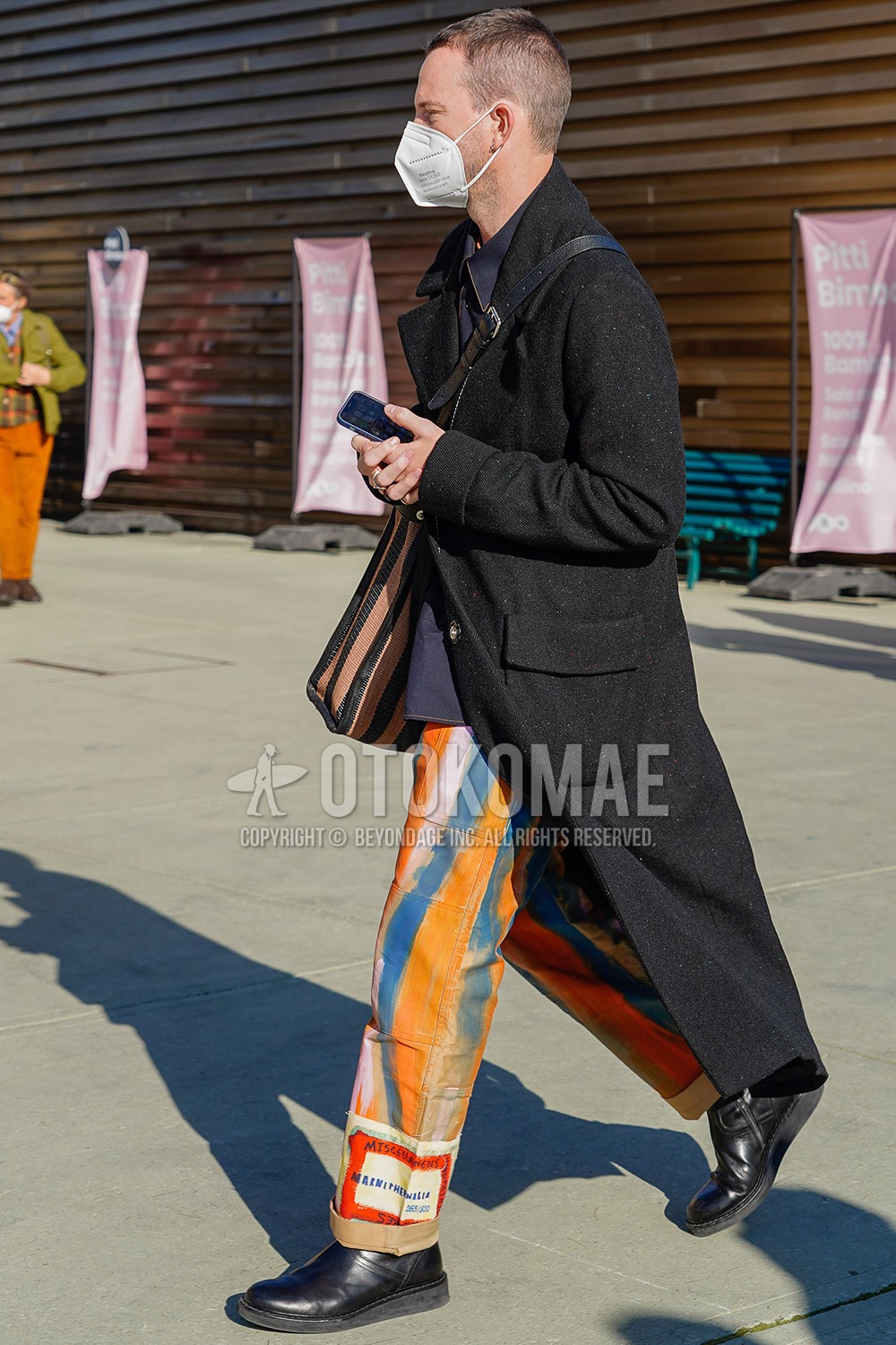 Men's spring winter outfit with black plain chester coat, navy plain shirt, orange blue stripes wide pants, black boots, brown black stripes shoulder bag.