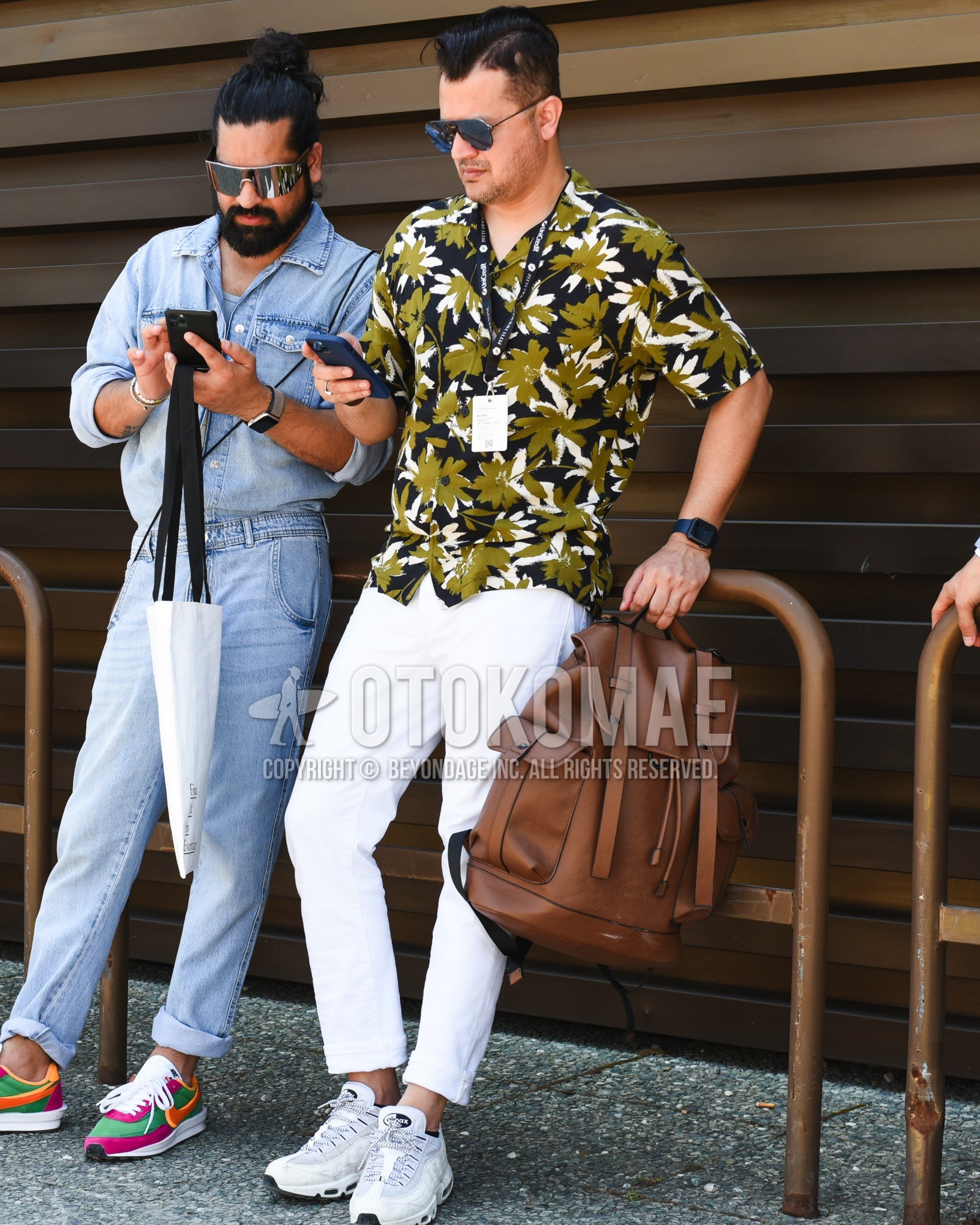 Men's spring summer outfit with black plain sunglasses, olive green black graphic shirt, white plain cotton pants, white plain cropped pants, white low-cut sneakers, brown plain backpack.