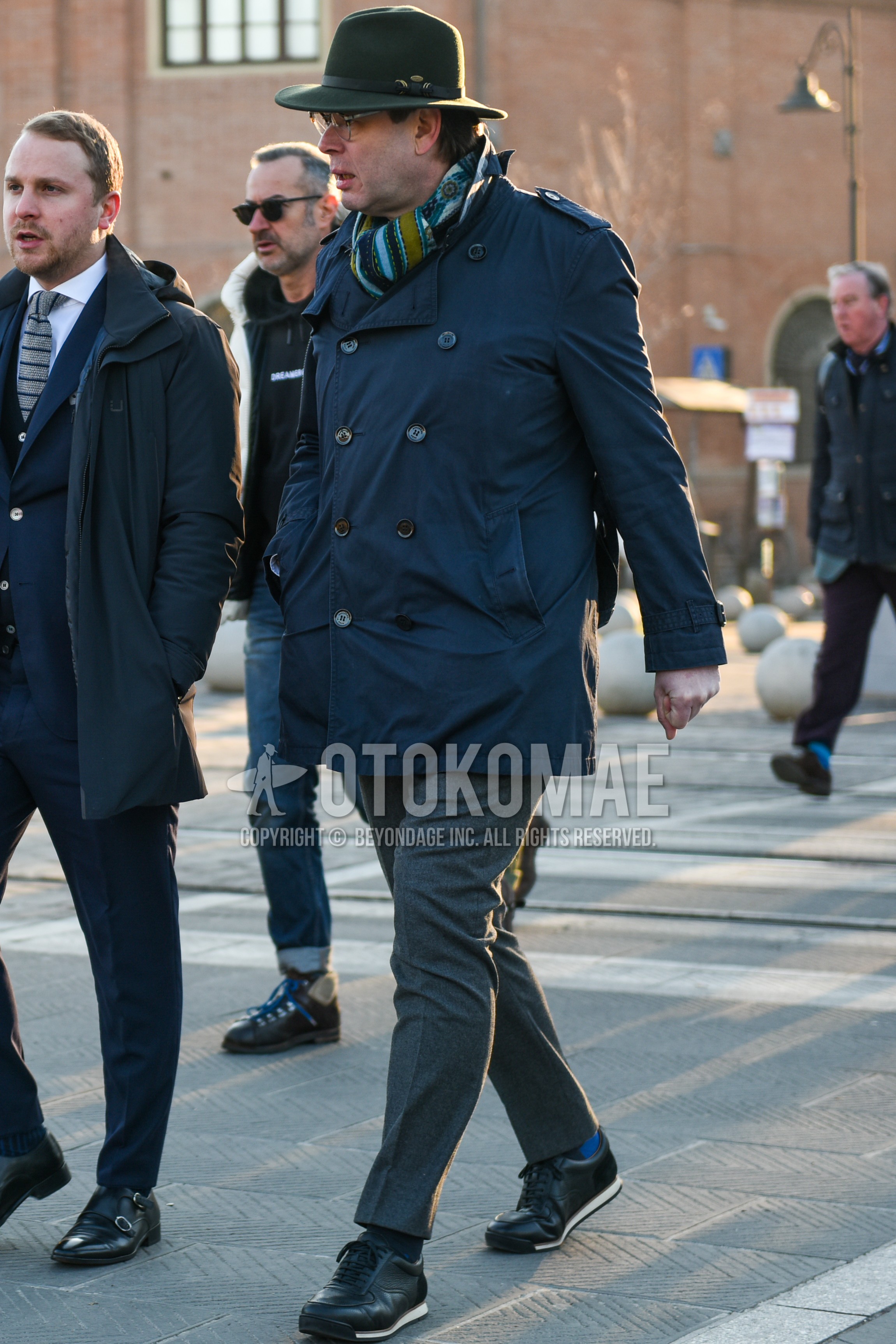 Men's autumn winter outfit with olive green plain hat, silver plain glasses, multi-color scarf scarf, navy plain trench coat, gray plain slacks, gray plain ankle pants, gray blue plain socks, black low-cut sneakers.