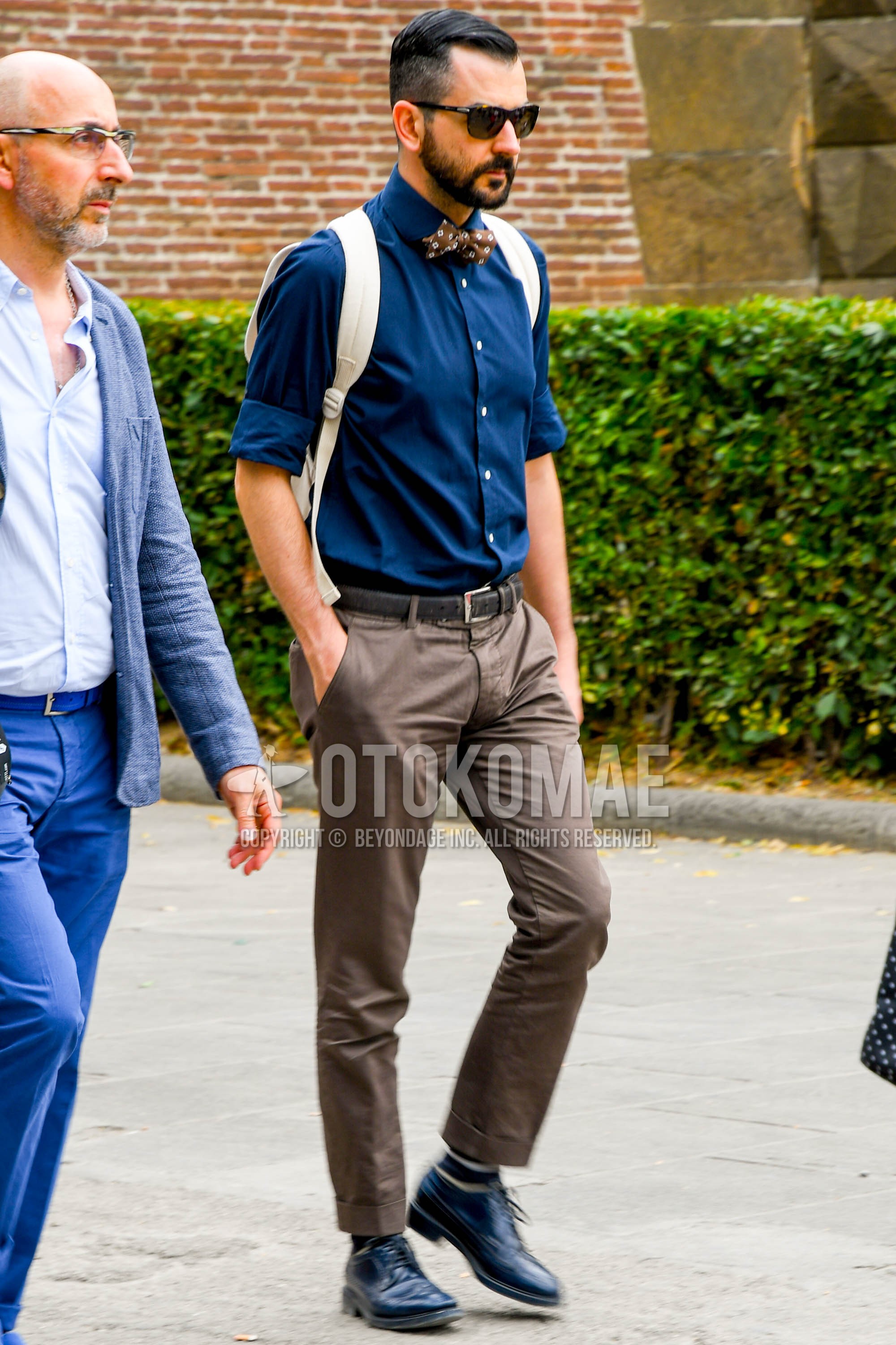 Men's spring summer outfit with brown tortoiseshell sunglasses, navy plain shirt, black plain leather belt, brown plain chinos, black horizontal stripes socks, black wing-tip shoes leather shoes, white plain backpack, brown necktie bow tie.