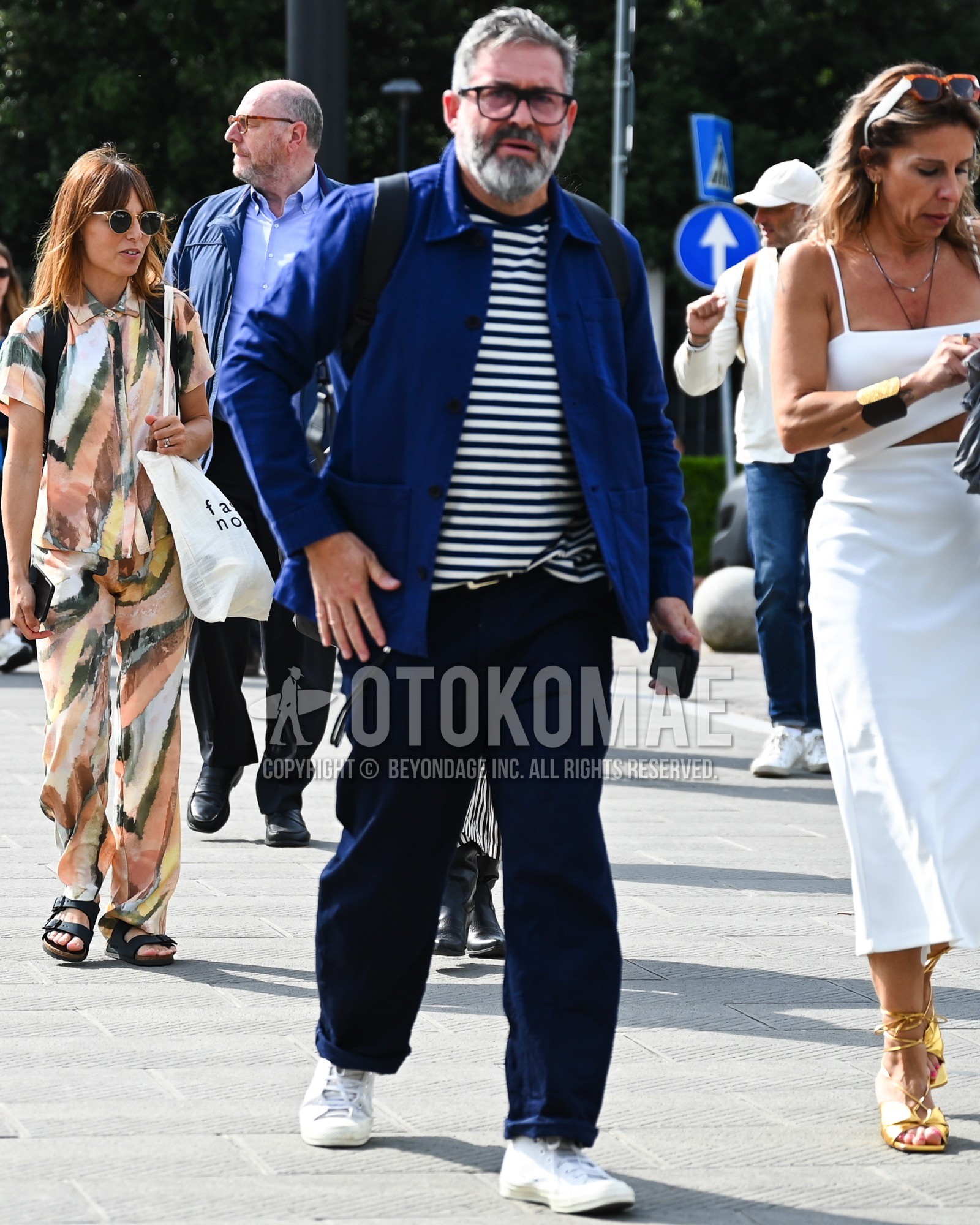 Men's spring summer autumn outfit with clear tortoiseshell sunglasses, blue plain coverall, white navy horizontal stripes t-shirt, navy plain chinos, white low-cut sneakers, black plain backpack.