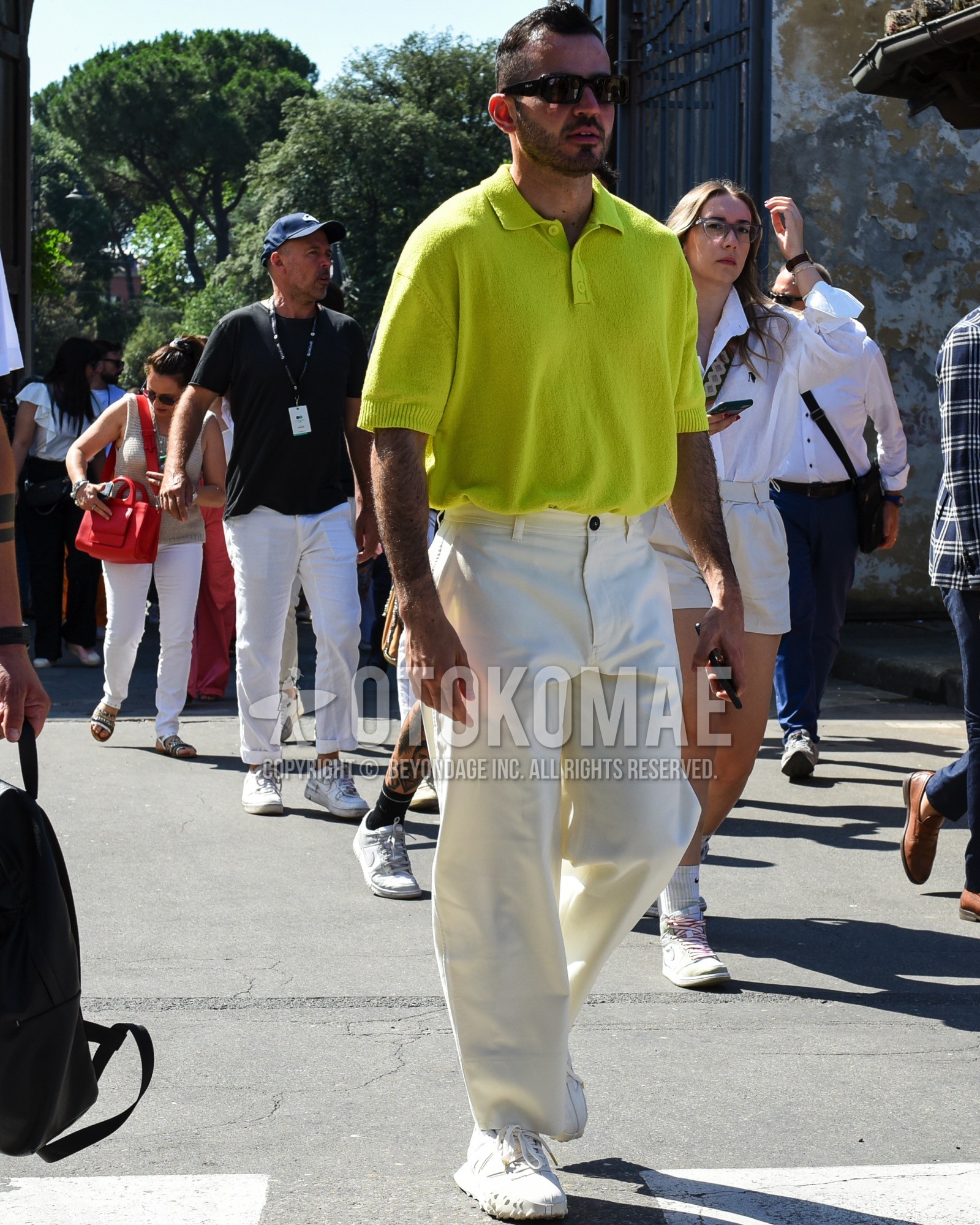 Men's spring summer outfit with black plain sunglasses, yellow plain polo shirt, yellow plain sweater, white plain cotton pants, white low-cut sneakers.
