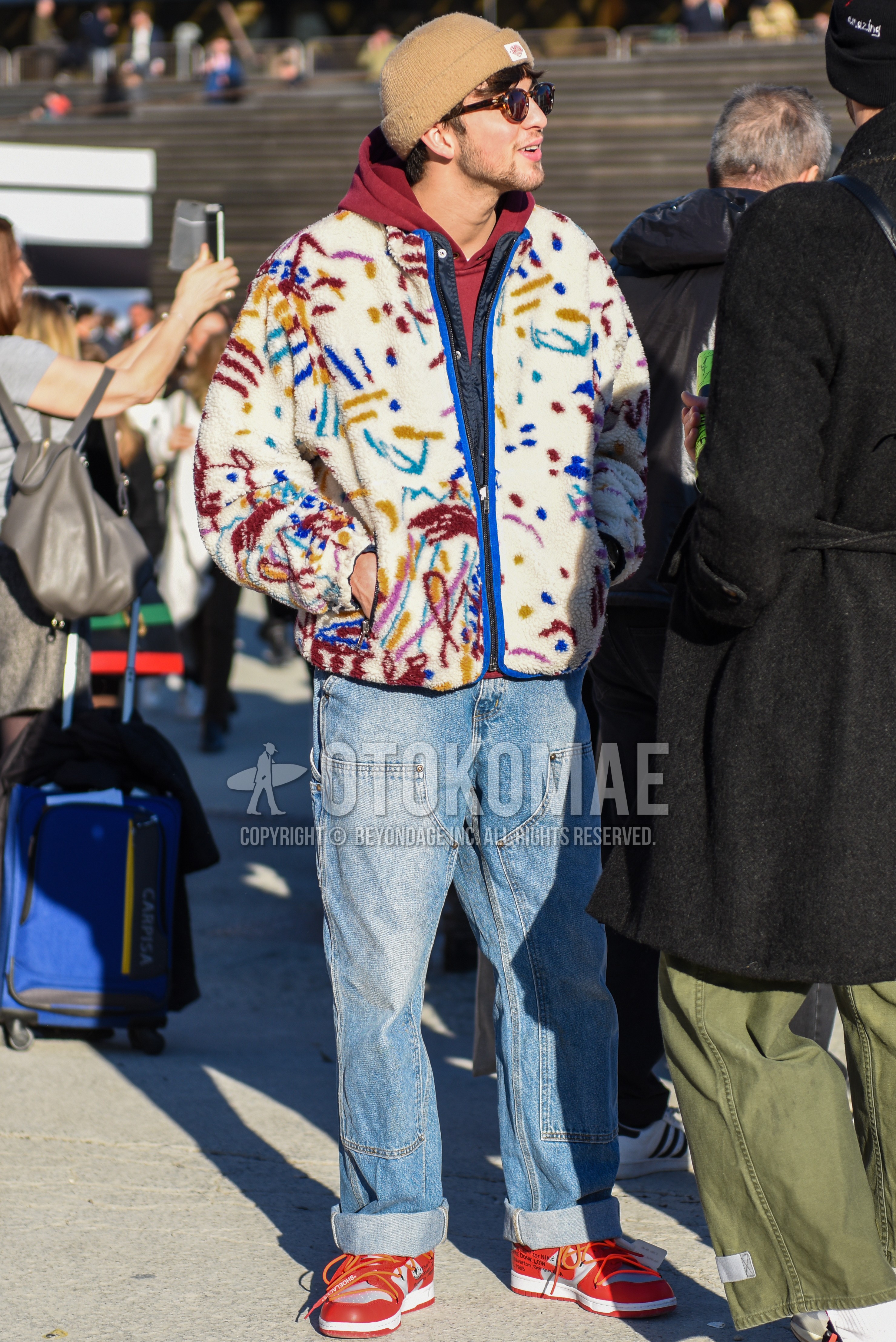 Men's autumn winter outfit with beige plain knit cap, brown tortoiseshell sunglasses, white graphic fleece jacket, red plain hoodie, blue plain denim/jeans, red low-cut sneakers.