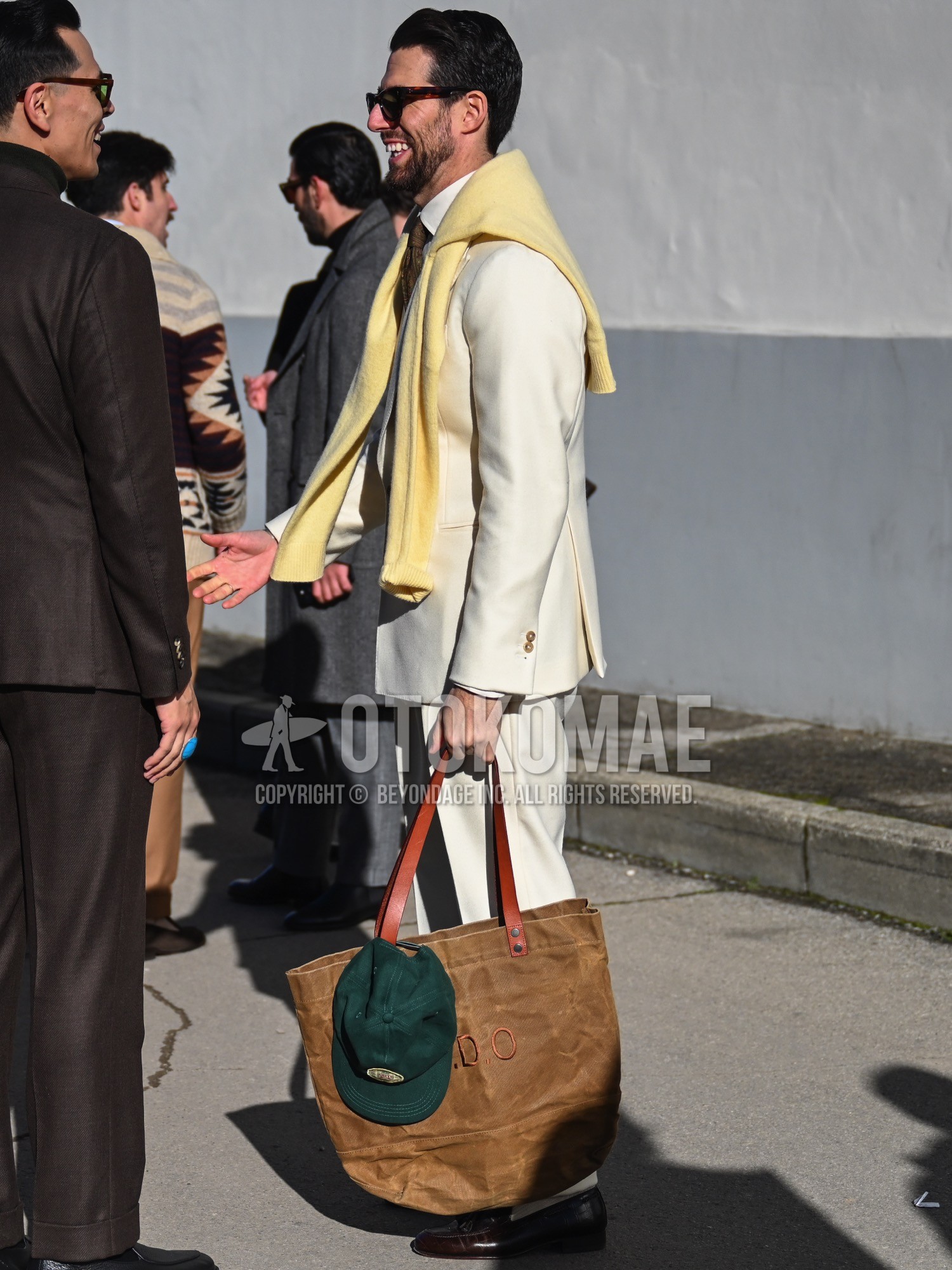 Men's spring autumn winter outfit with green one point baseball cap, brown tortoiseshell sunglasses, white plain shirt, yellow plain cardigan, brown tassel loafers leather shoes, brown plain tote bag, white plain suit, brown paisley necktie.