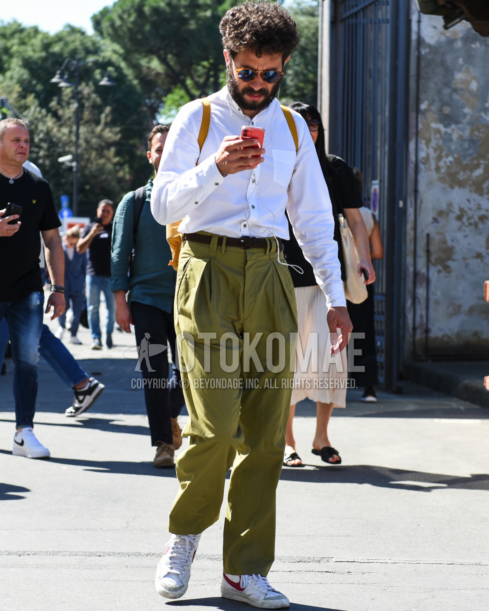 Men's spring summer outfit with brown tortoiseshell sunglasses, white plain shirt, brown plain braided belt, olive green plain chinos, white low-cut sneakers, yellow plain backpack.