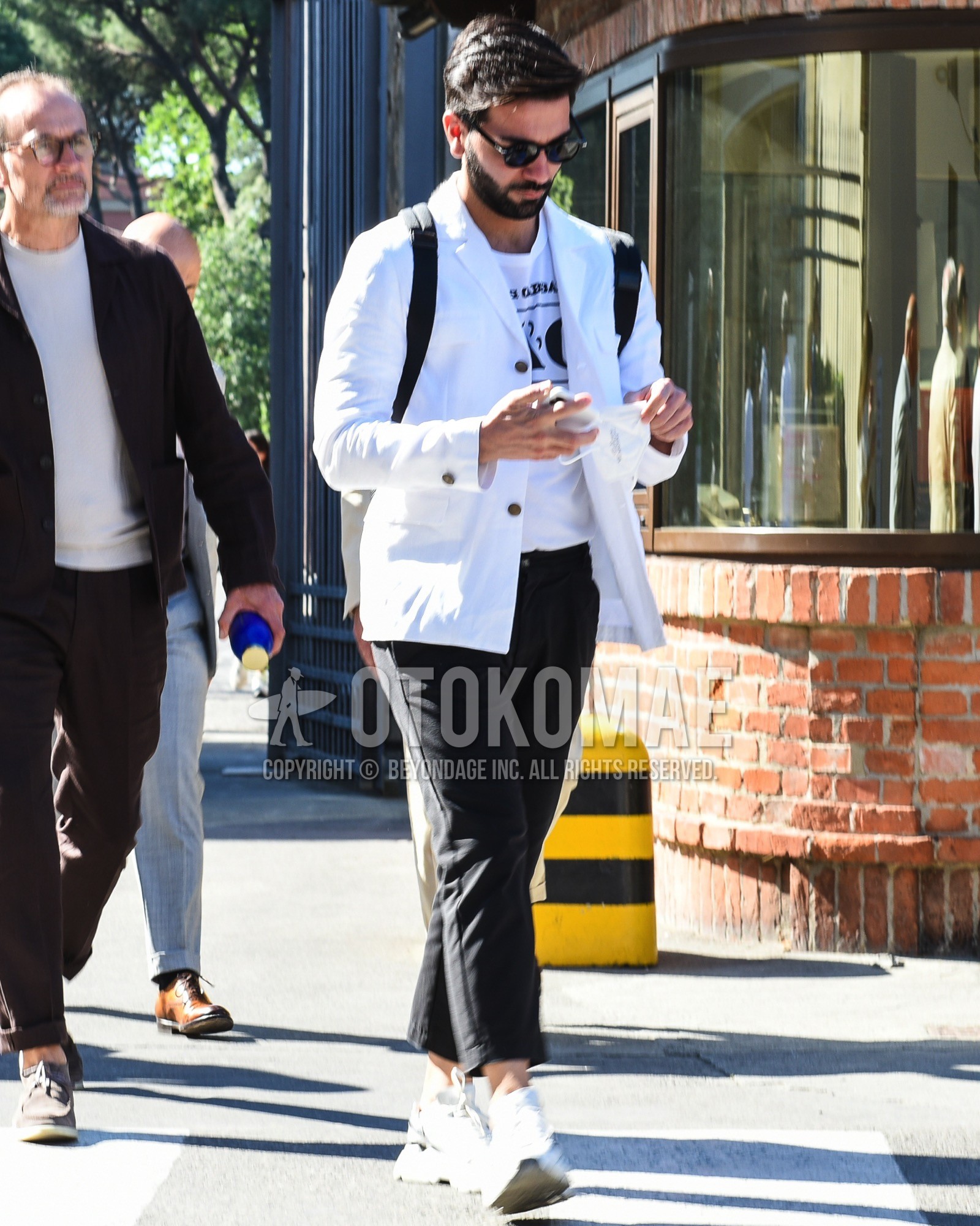 Men's spring summer outfit with black plain sunglasses, white plain tailored jacket, white deca logo t-shirt, black plain chinos, black plain cropped pants, white low-cut sneakers, black plain backpack.