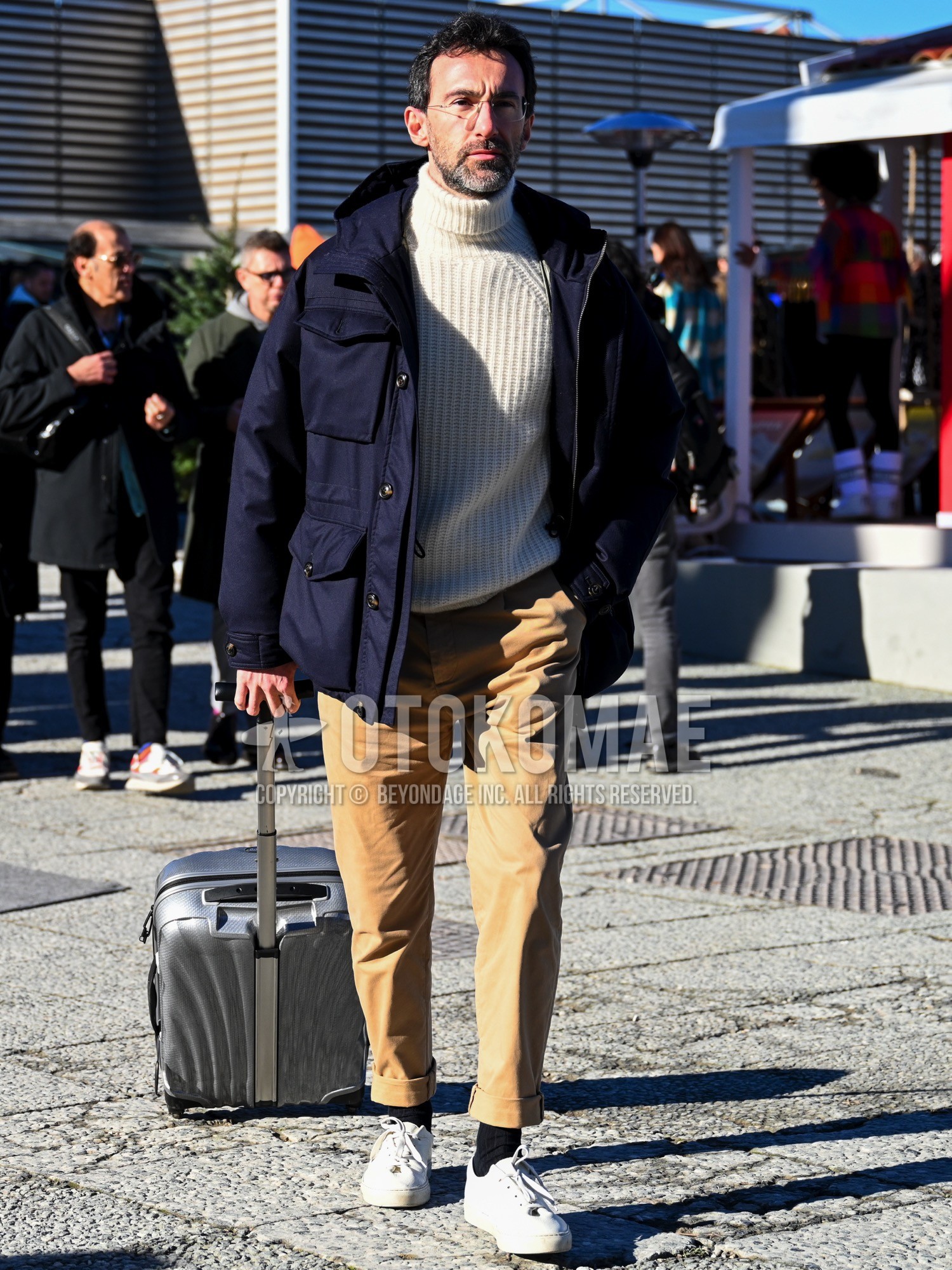 Men's autumn winter outfit with navy plain hooded coat, navy plain military jacket, white plain turtleneck knit, beige plain chinos, black plain socks, white low-cut sneakers, gray plain suitcase.