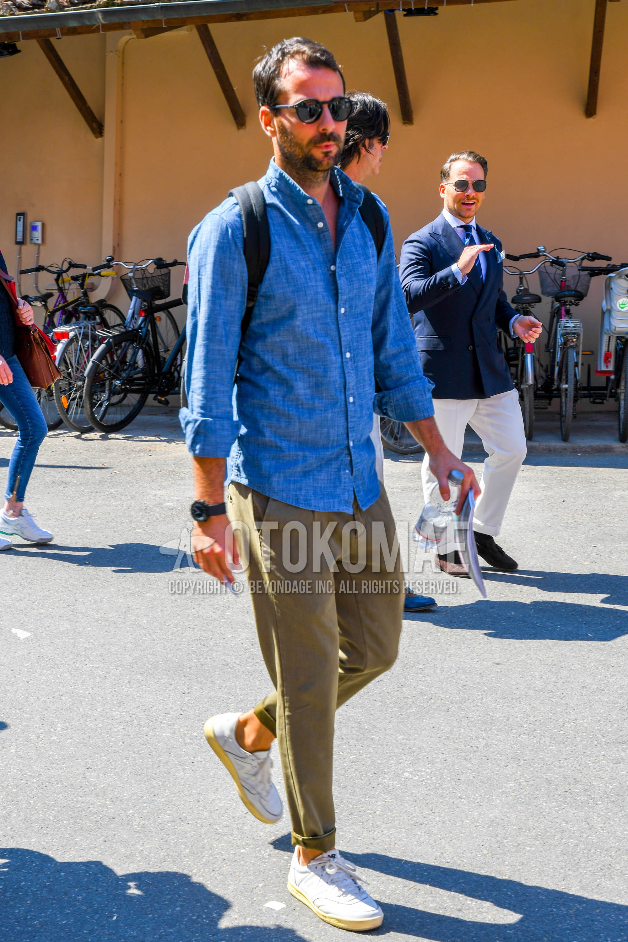 Men's spring summer outfit with plain sunglasses, blue plain denim shirt/chambray shirt, beige plain chinos, white low-cut sneakers.