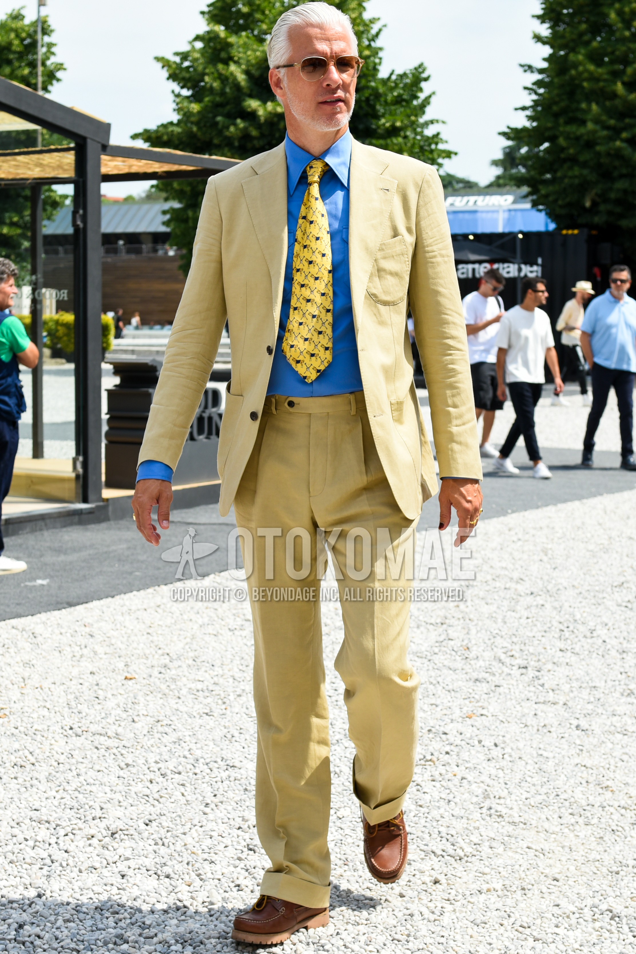 Men's spring summer outfit with black plain sunglasses, blue plain shirt, brown moccasins/deck shoes leather shoes, yellow beige plain suit, yellow small crest necktie.