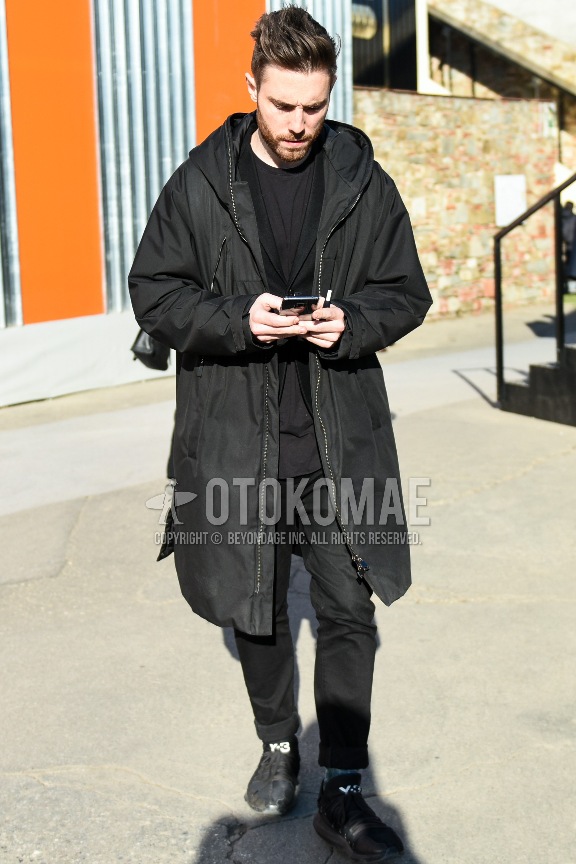 Men's autumn winter outfit with black plain hooded coat, black plain t-shirt, black plain tailored jacket, black plain slacks, black low-cut sneakers.