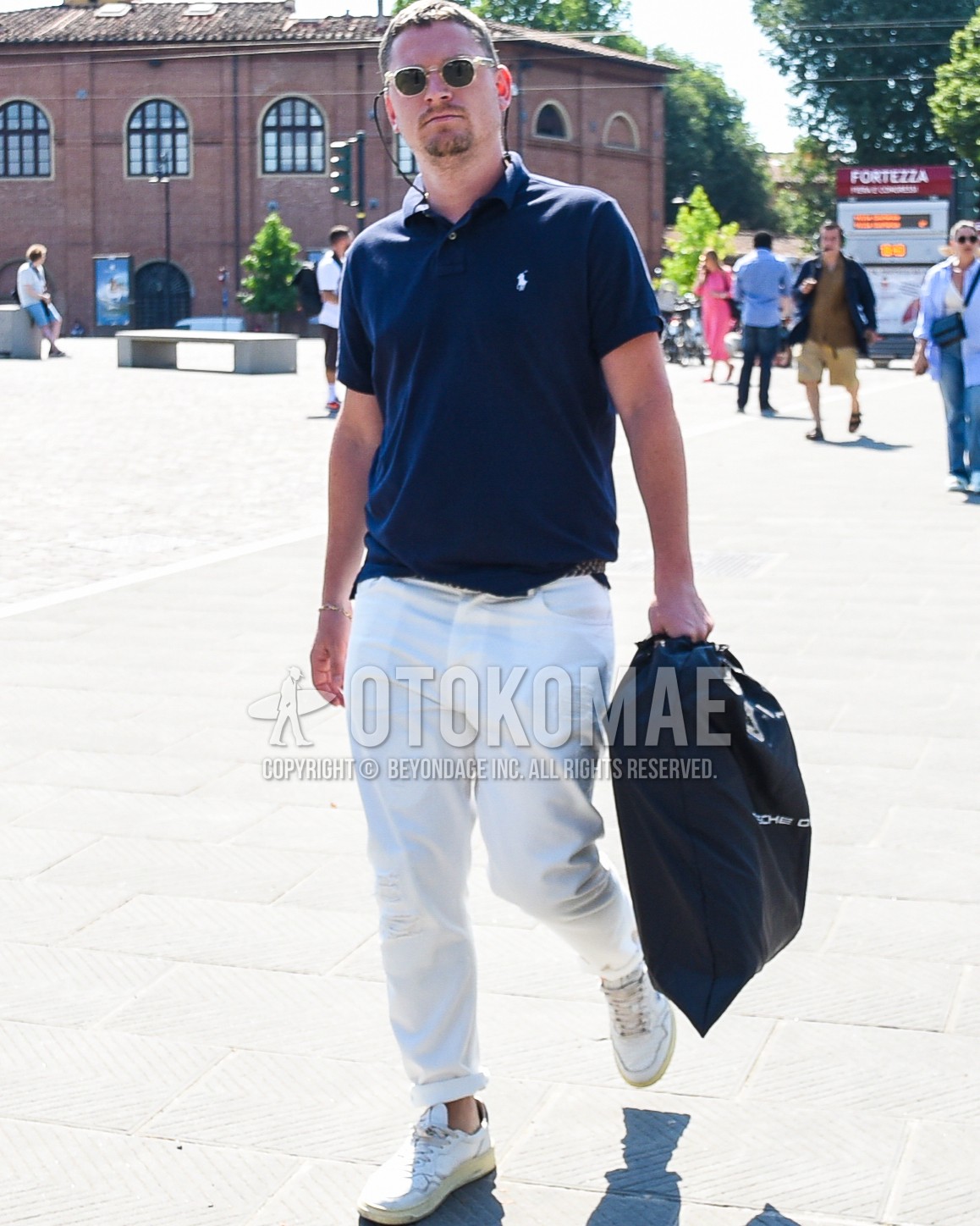 Men's spring summer outfit with black plain sunglasses, navy one point polo shirt, white plain chinos, white low-cut sneakers, black deca logo tote bag.