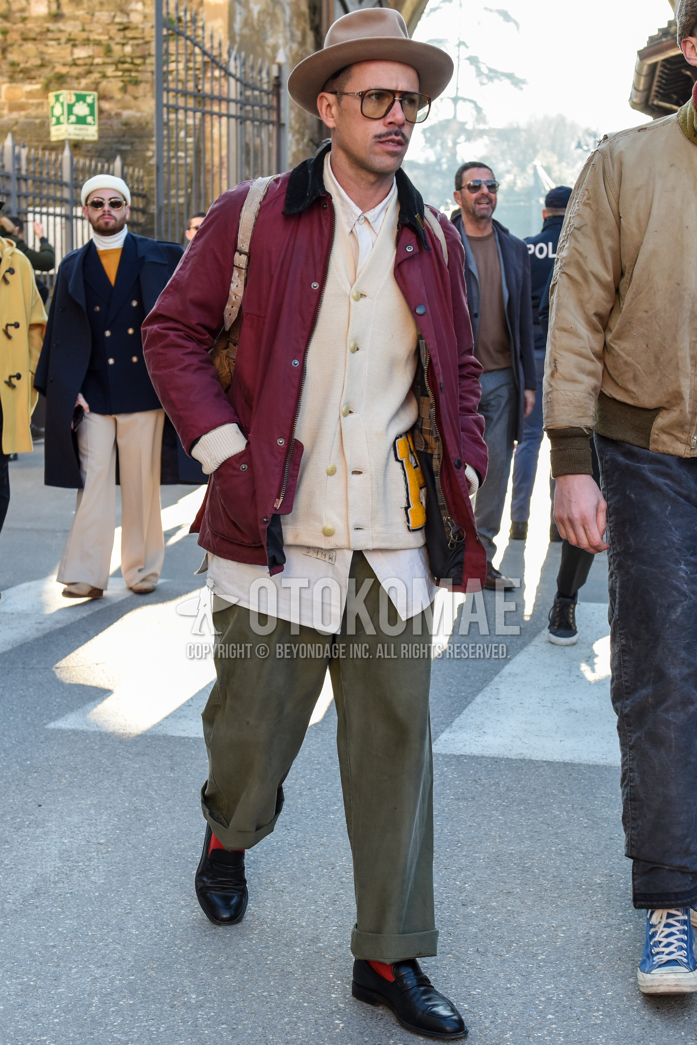 Men's autumn winter outfit with beige plain hat, brown tortoiseshell sunglasses, red plain field jacket/hunting jacket, white lettered cardigan, white plain shirt, olive green plain chinos, orange plain socks, black coin loafers leather shoes.