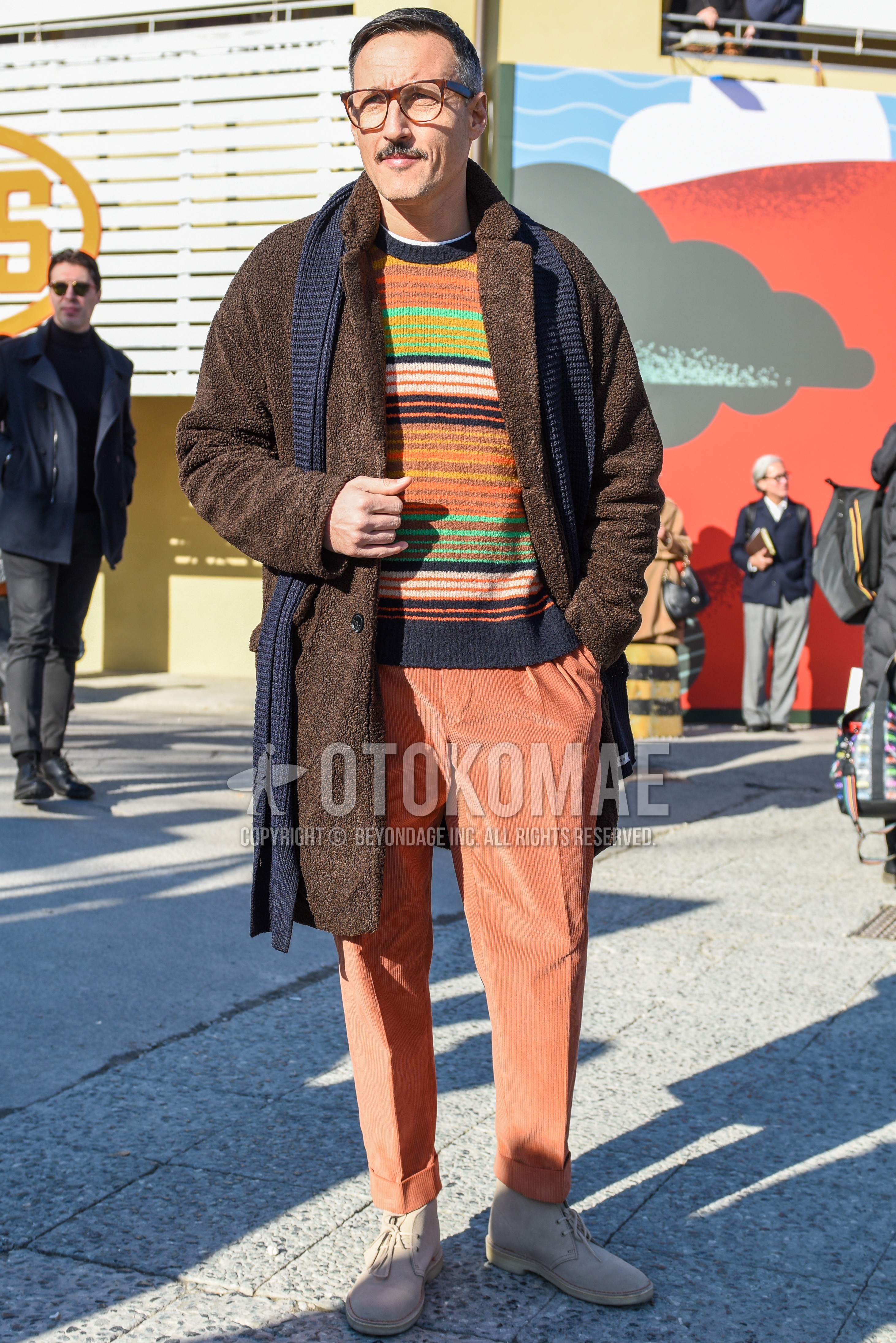 Men's autumn winter outfit with brown tortoiseshell glasses, brown plain chester coat, multi-color horizontal stripes sweater, orange plain winter pants (corduroy,velour), beige chukka boots.