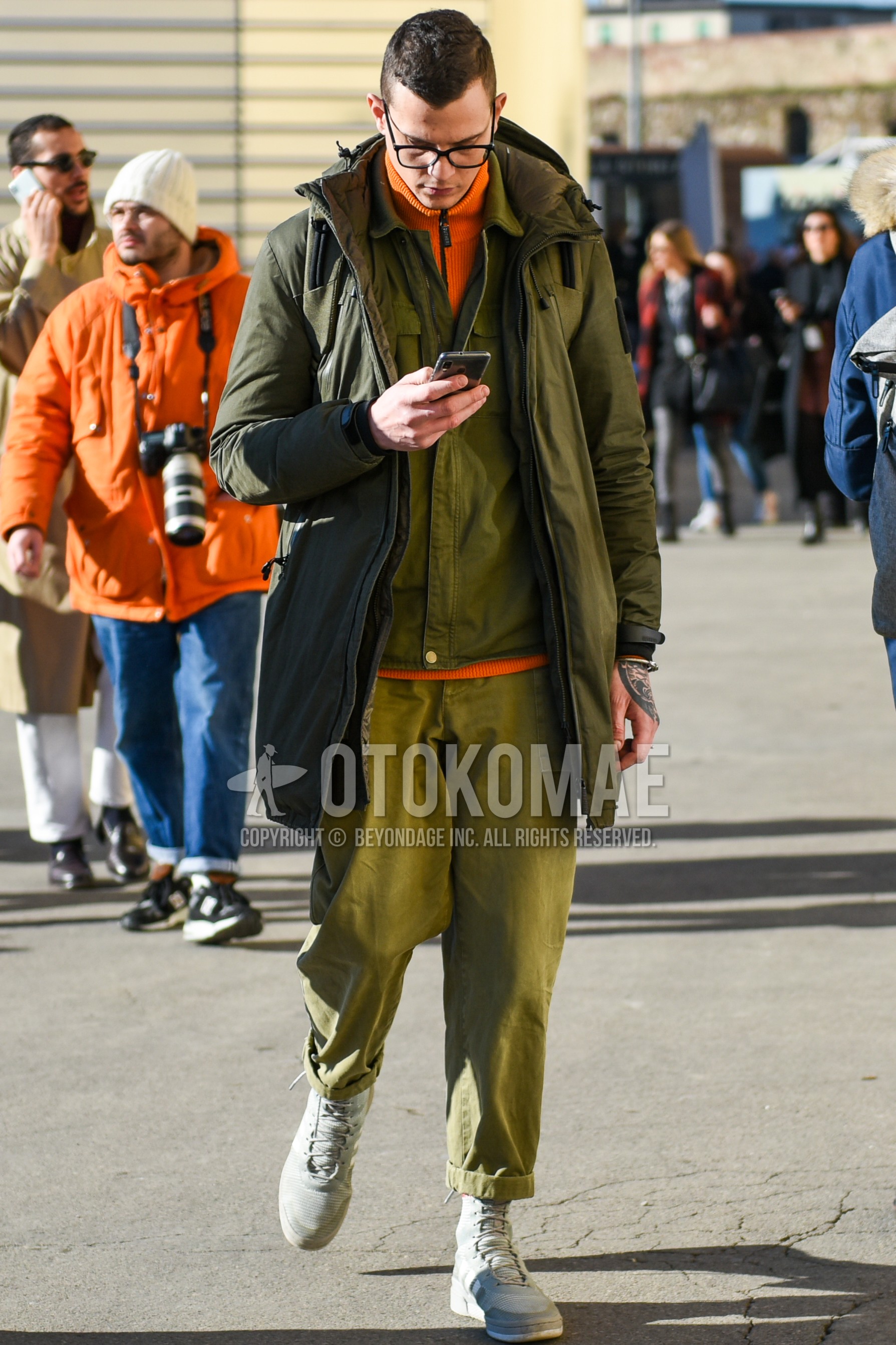 Men's winter outfit with black plain glasses, olive green plain hooded coat, olive green plain shirt jacket, orange plain cardigan, olive green plain chinos, gray high-cut sneakers.