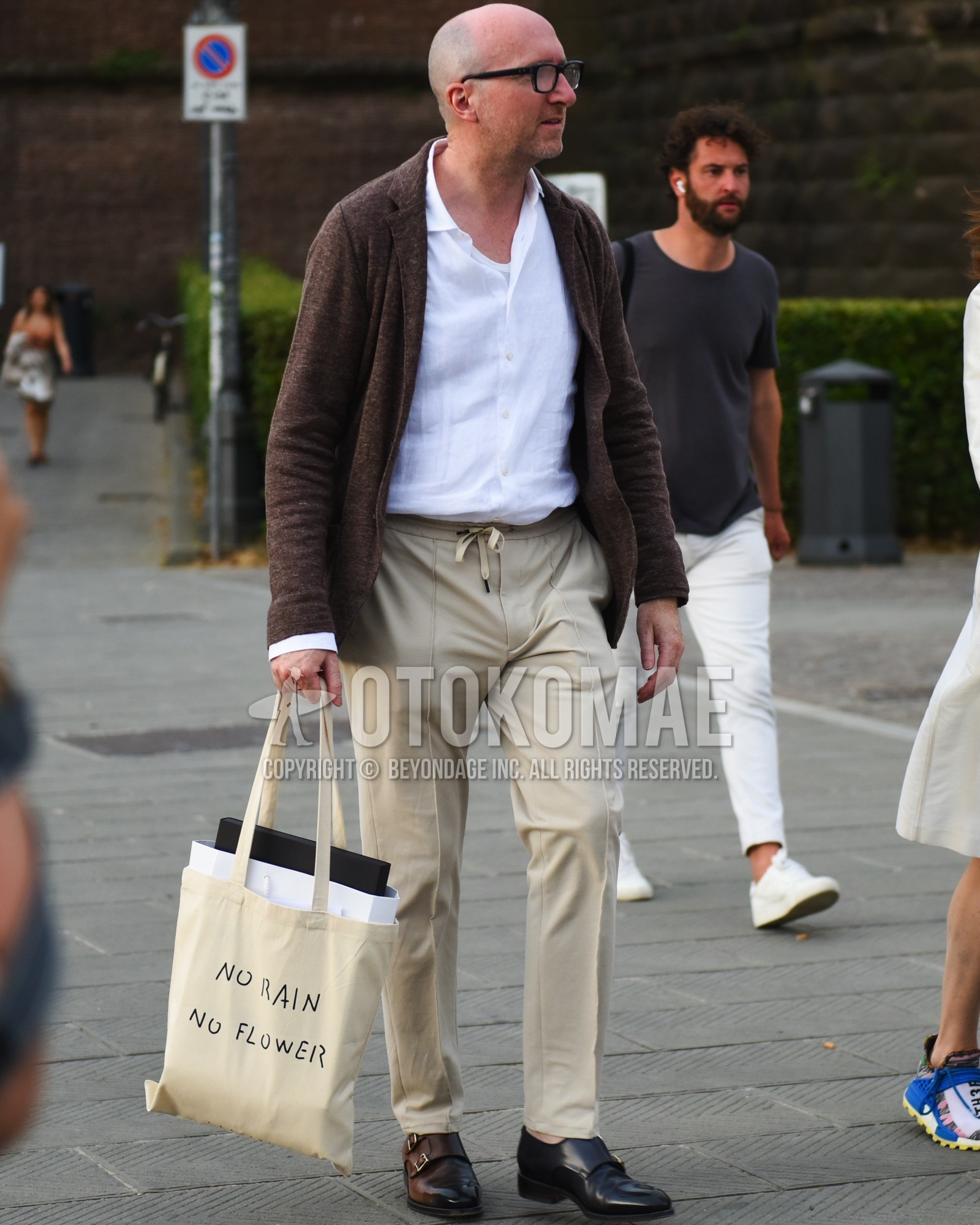 Men's spring summer outfit with black plain glasses, brown plain tailored jacket, white plain shirt, beige plain easy pants, brown monk shoes leather shoes, white deca logo tote bag.