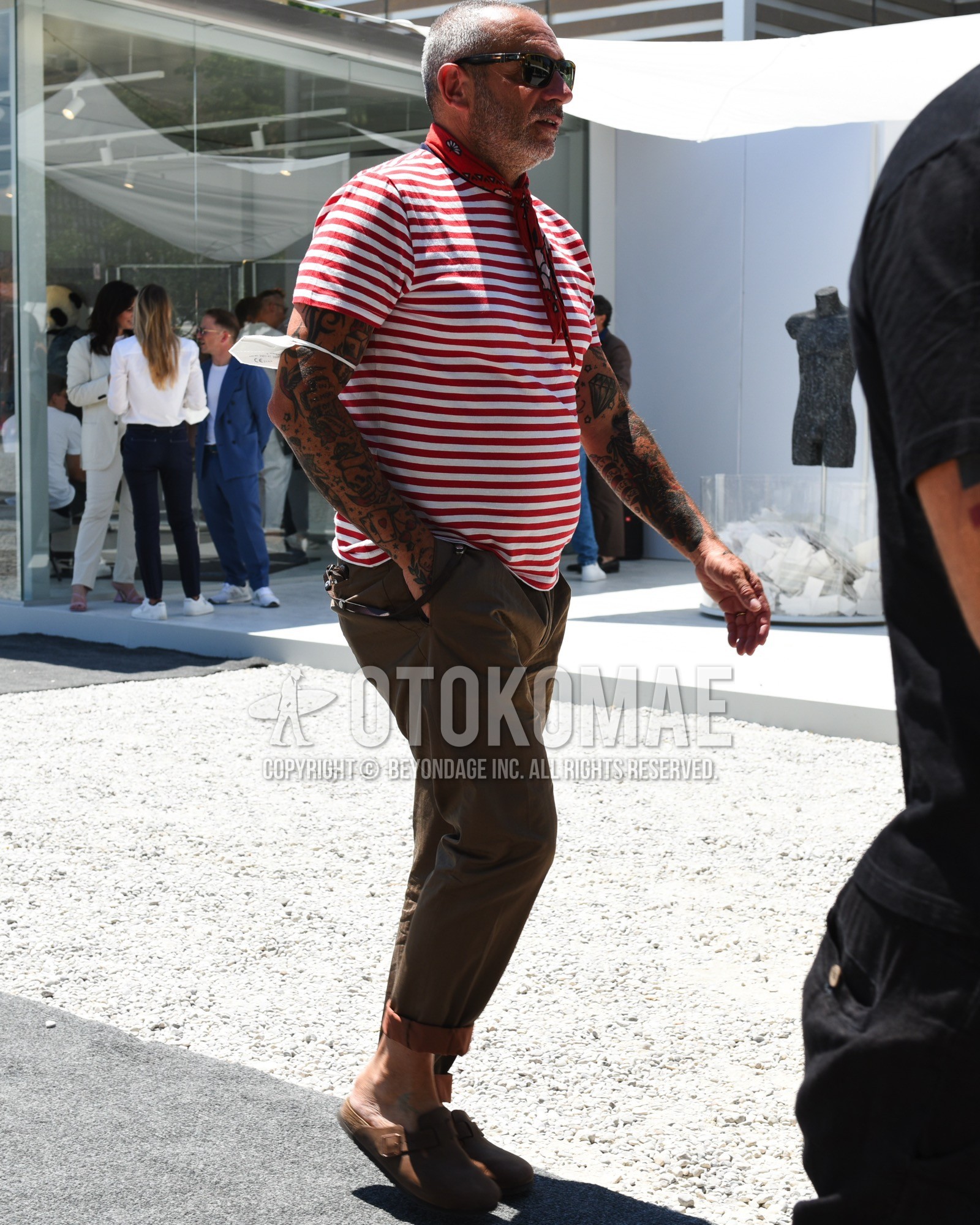 Men's spring summer outfit with black plain sunglasses, red scarf bandana/neckerchief, red white horizontal stripes t-shirt, brown plain slacks, brown leather sandals.