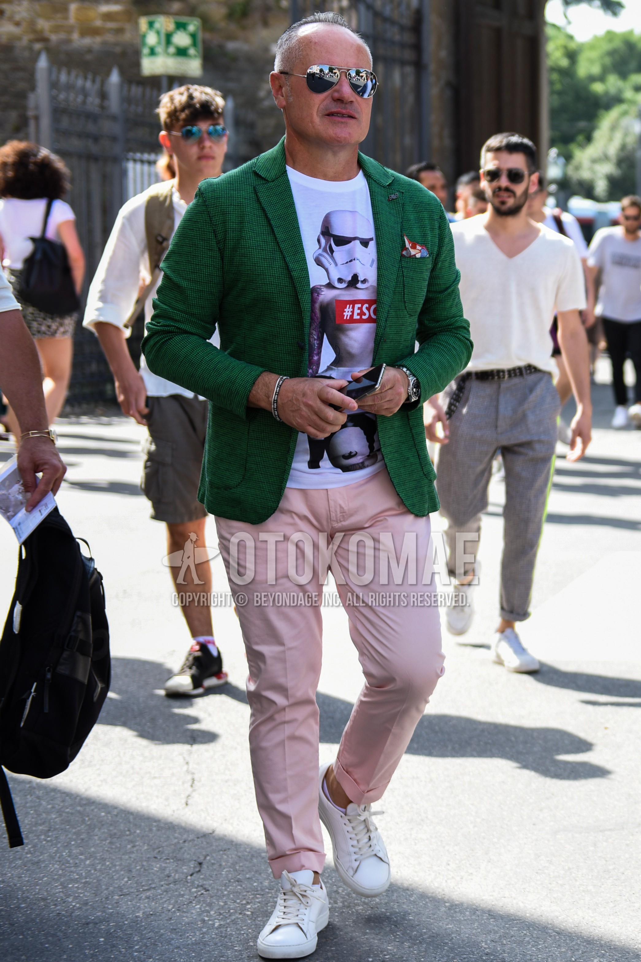 Men's spring summer outfit with silver plain sunglasses, green plain tailored jacket, white graphic t-shirt, pink plain chinos, white low-cut sneakers.