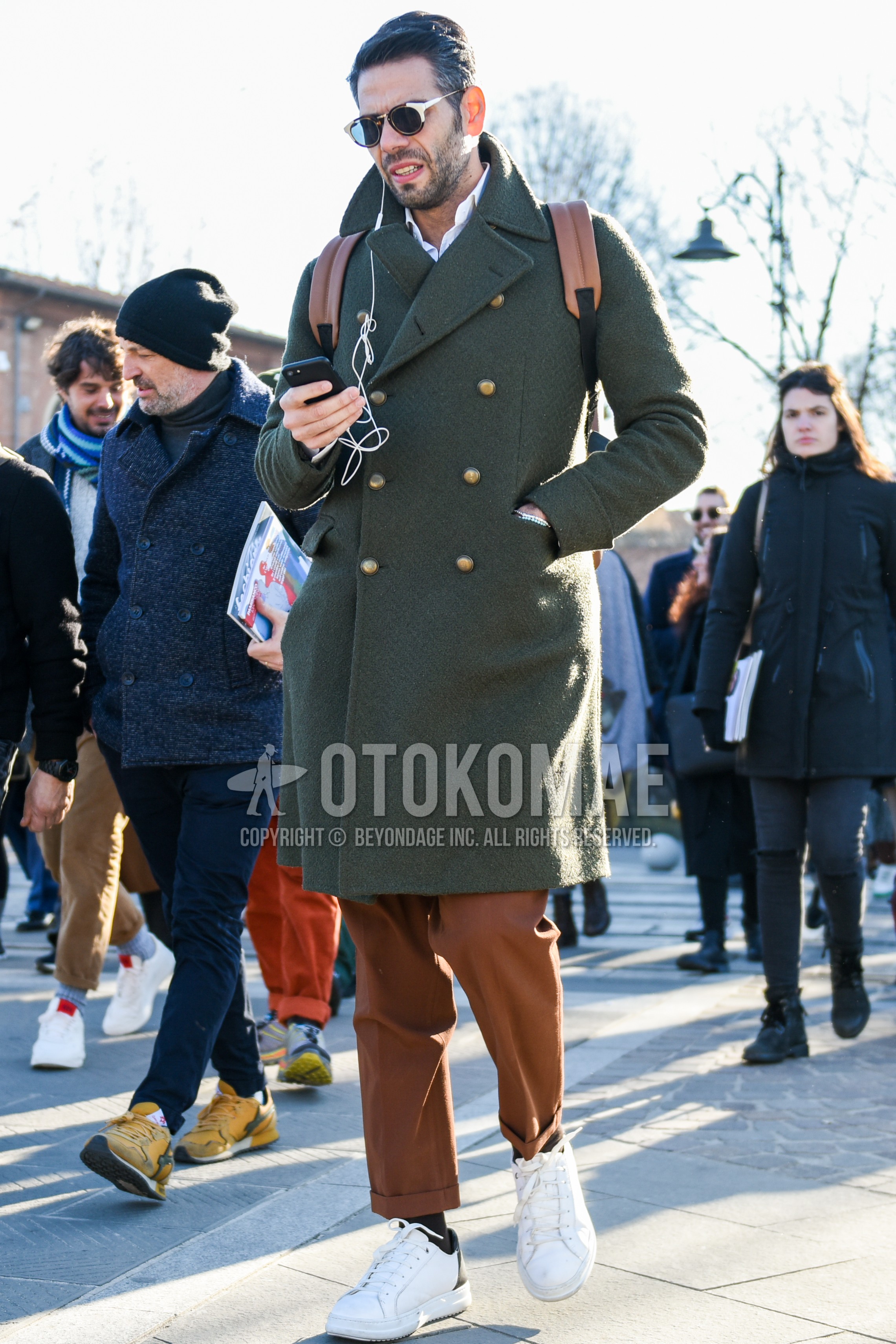 Men's autumn winter outfit with brown tortoiseshell sunglasses, olive green plain ulster coat, white plain shirt, brown plain slacks, brown plain ankle pants, black plain socks, white low-cut sneakers.