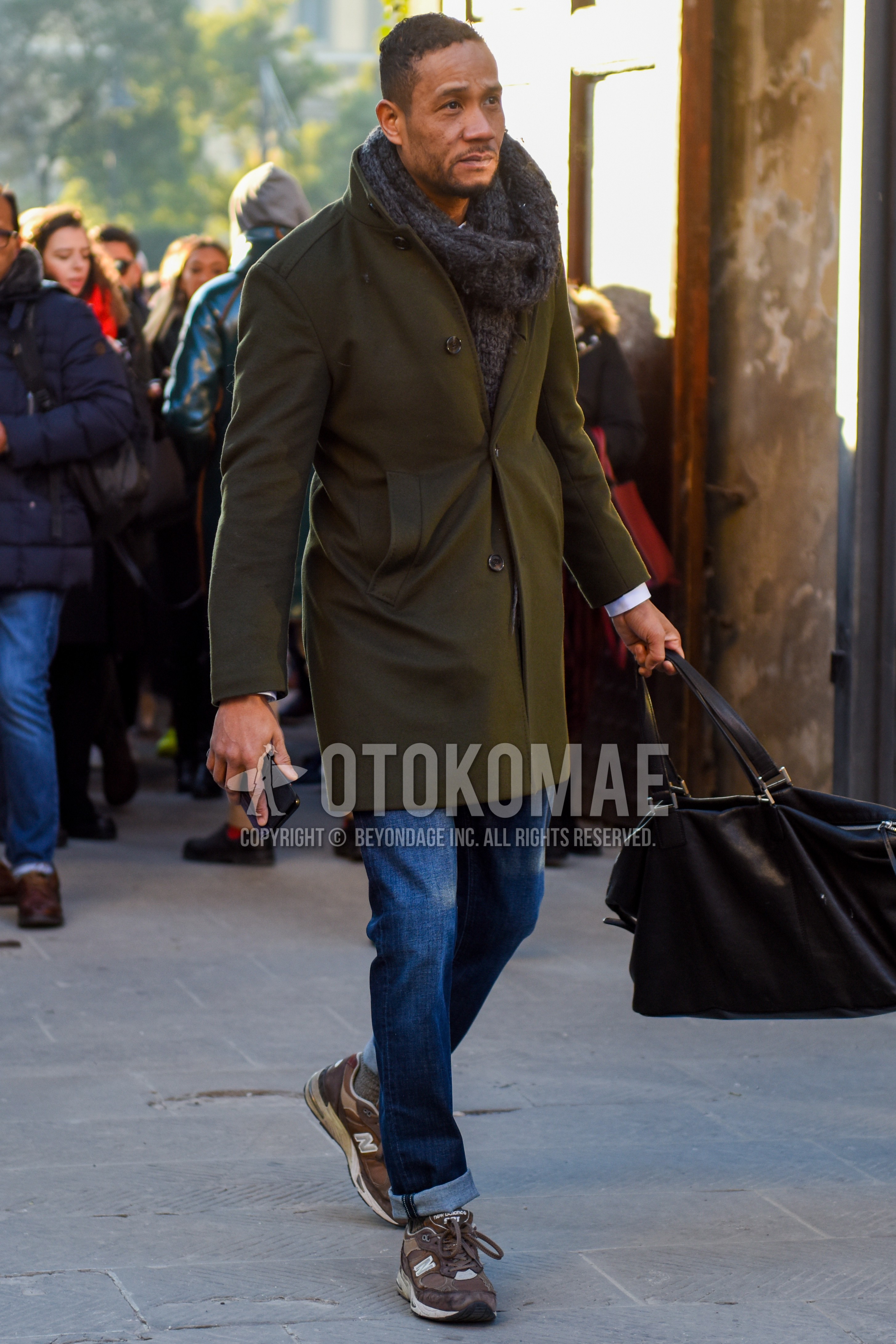 Men's winter outfit with gray plain scarf, olive green plain stenkarrer coat, blue plain denim/jeans, gray low-cut sneakers, black plain briefcase/handbag.