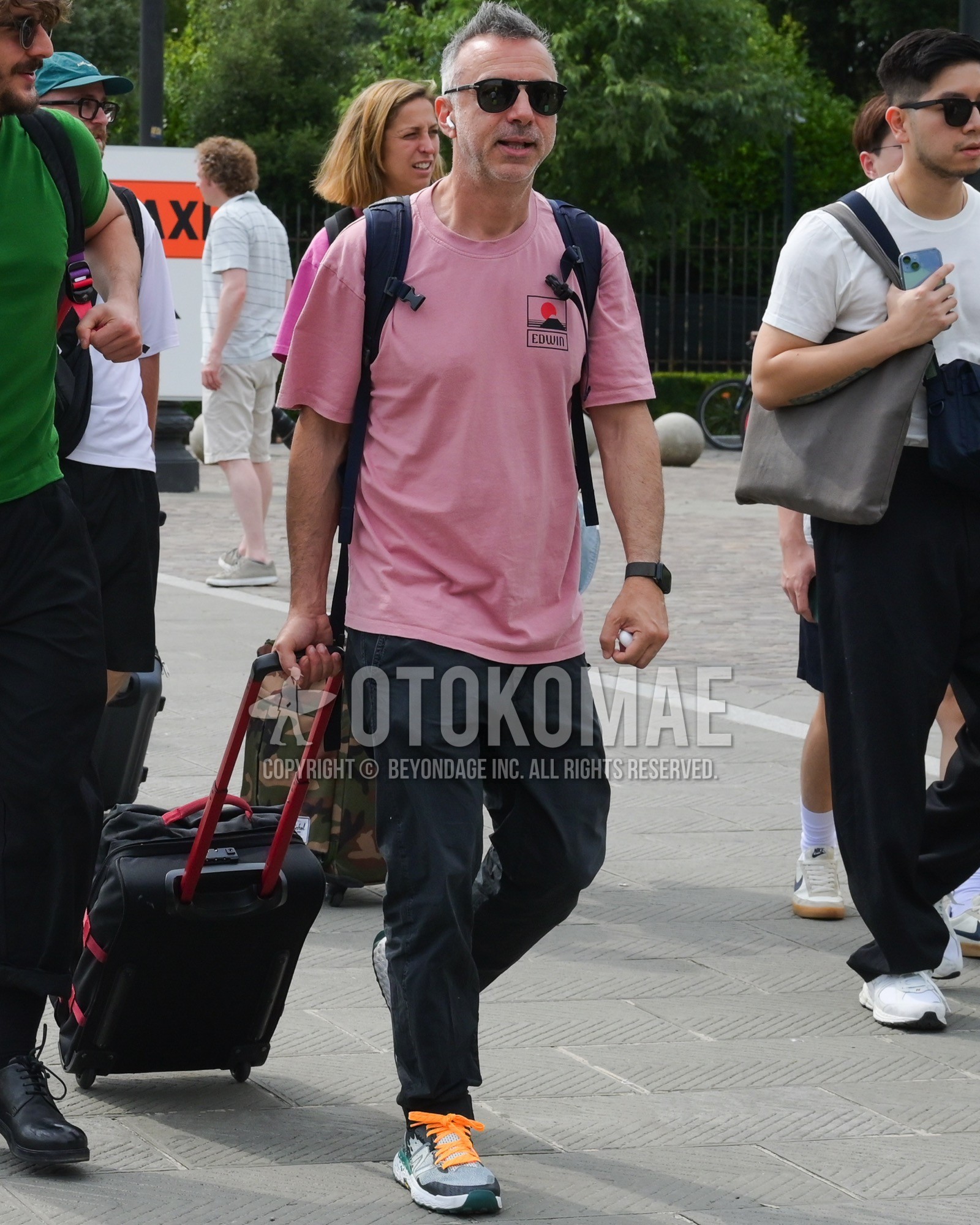 Men's spring summer outfit with black plain sunglasses, pink one point t-shirt, black plain chinos, gray low-cut sneakers, black plain backpack, black plain suitcase.