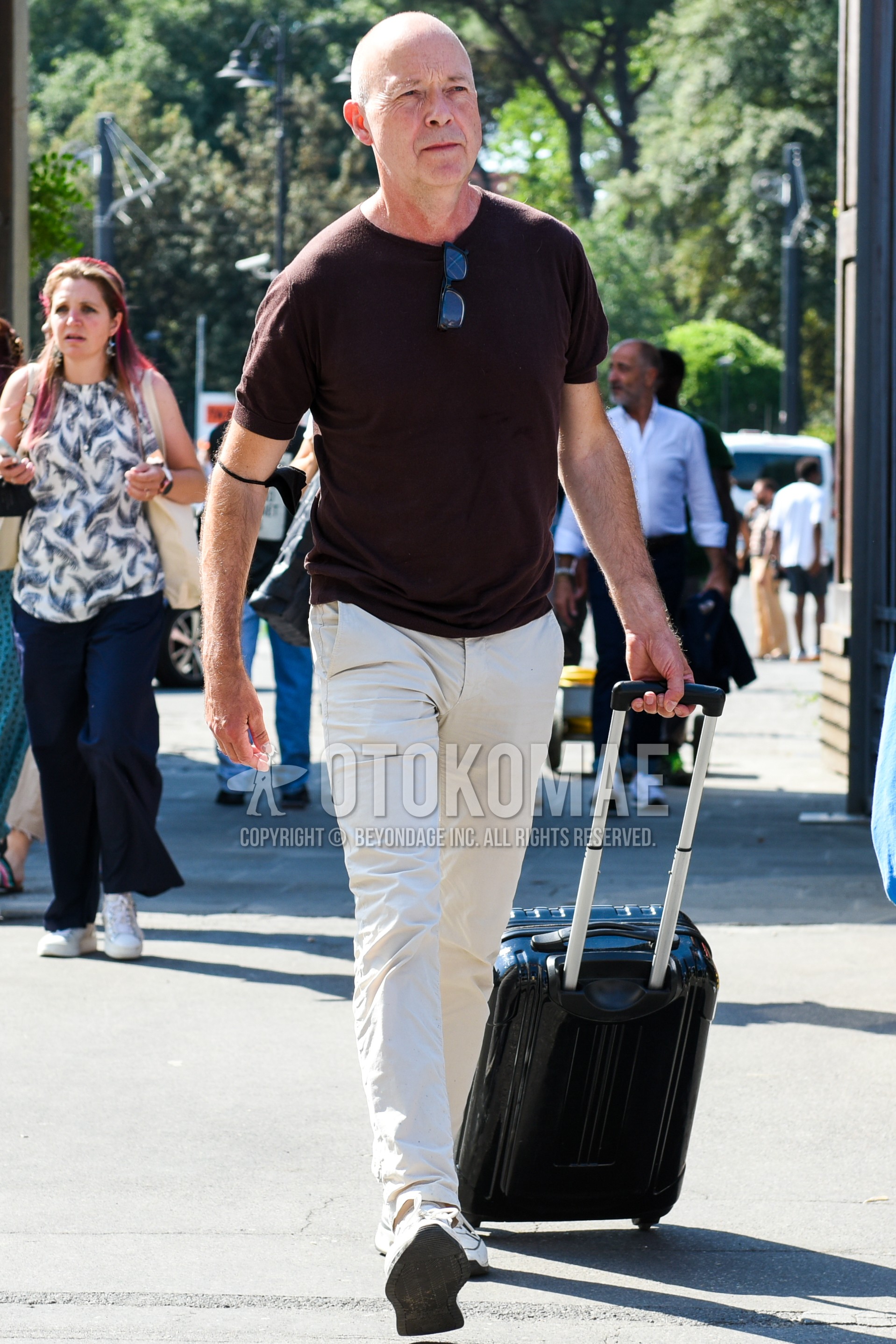 Men's spring summer outfit with brown plain t-shirt, beige plain chinos, white low-cut sneakers, black plain suitcase.
