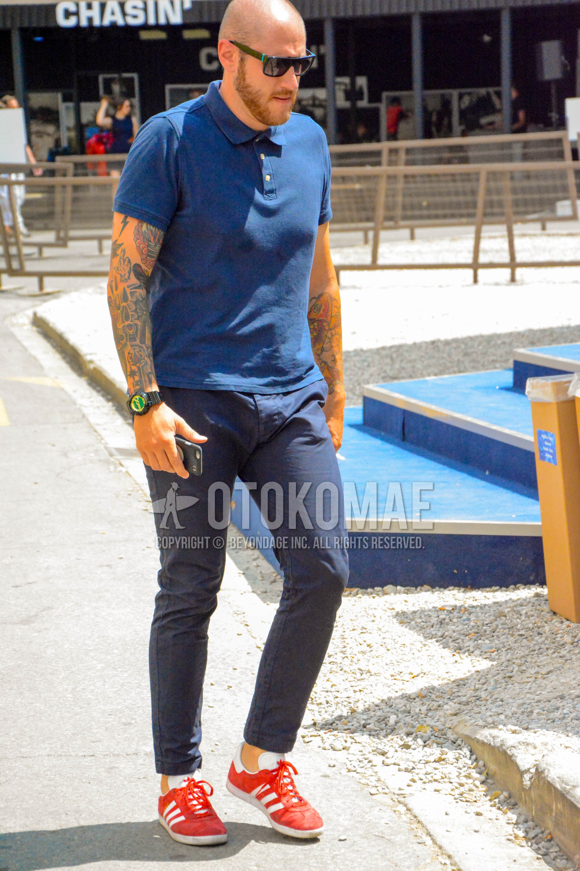 Men's spring summer outfit with black plain sunglasses, navy plain polo shirt, navy plain chinos, red low-cut sneakers.