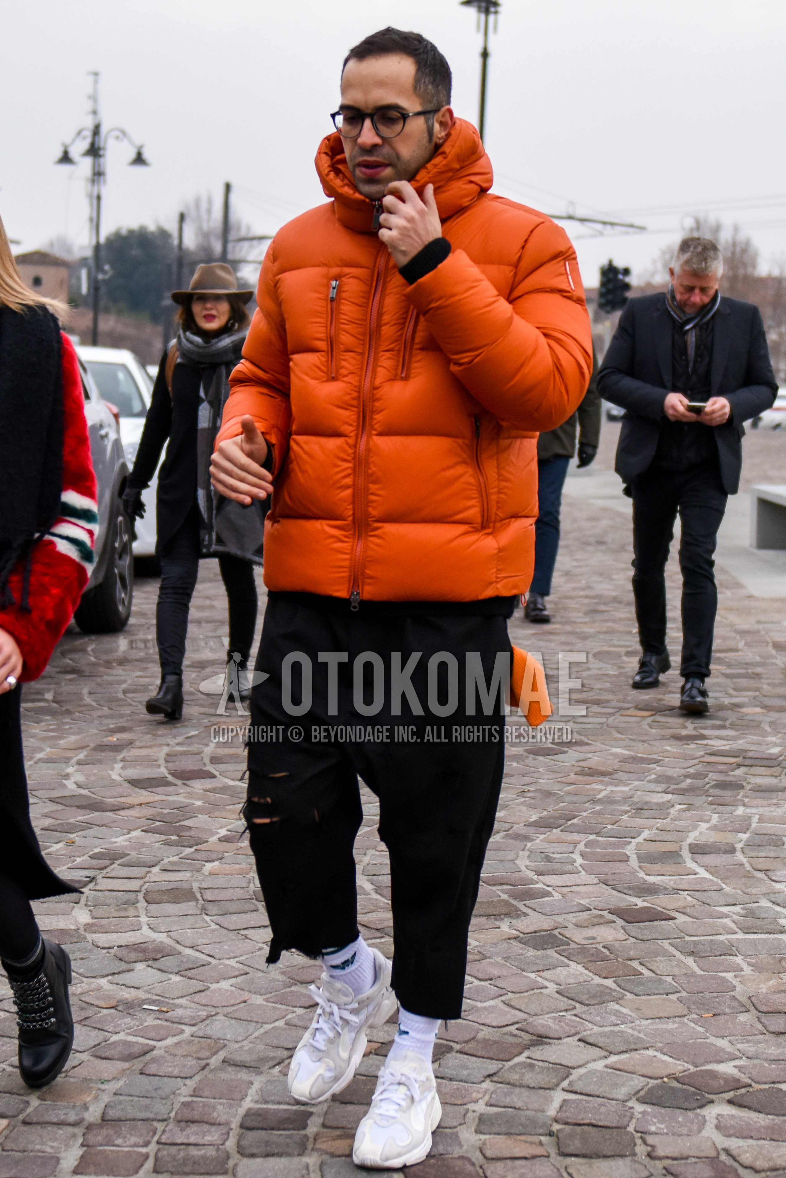 Men's winter outfit with plain glasses, orange plain down jacket, black plain damaged jeans, white plain socks, white low-cut sneakers.