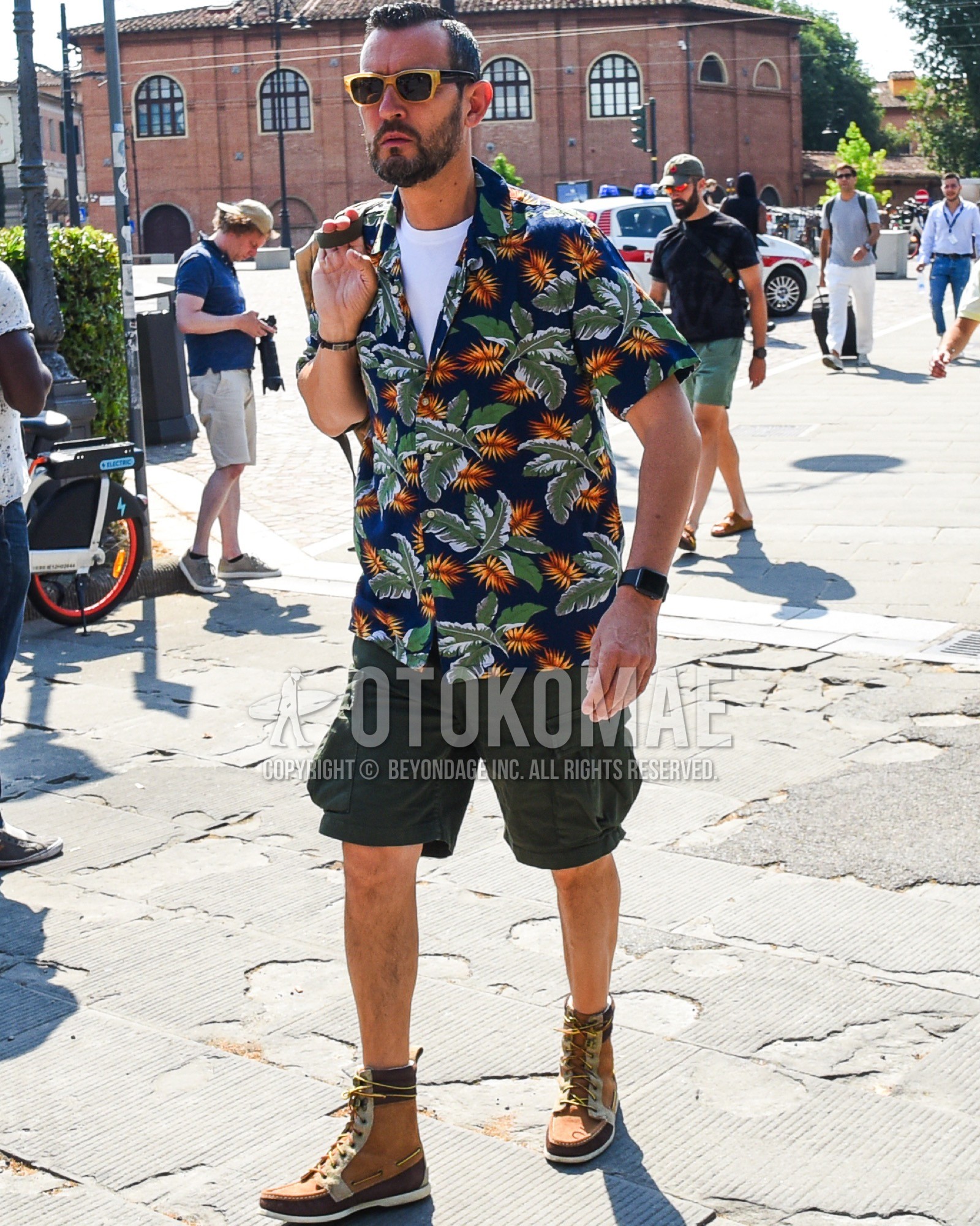 Men's spring summer outfit with yellow plain sunglasses, navy botanical shirt, white plain t-shirt, olive green plain cargo pants, olive green plain short pants, beige work boots.