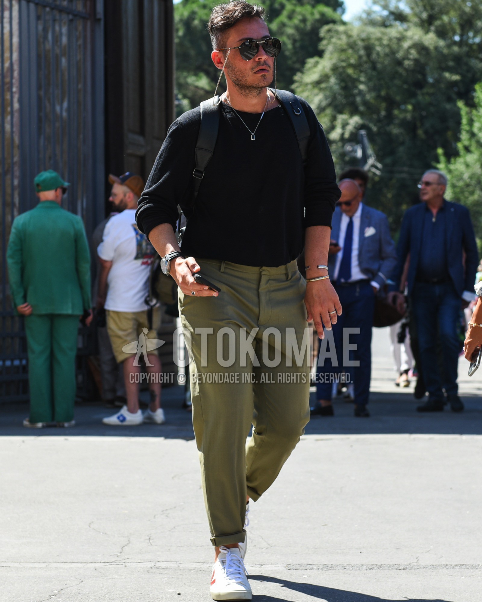 Men's spring summer outfit with black plain sunglasses, black plain long sleeve t-shirt, olive green plain chinos, white low-cut sneakers, black plain backpack.