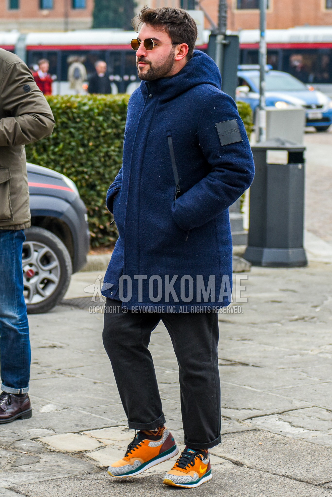 Men's winter outfit with gold plain sunglasses, navy plain hooded coat, gray plain chinos, multi-color low-cut sneakers.