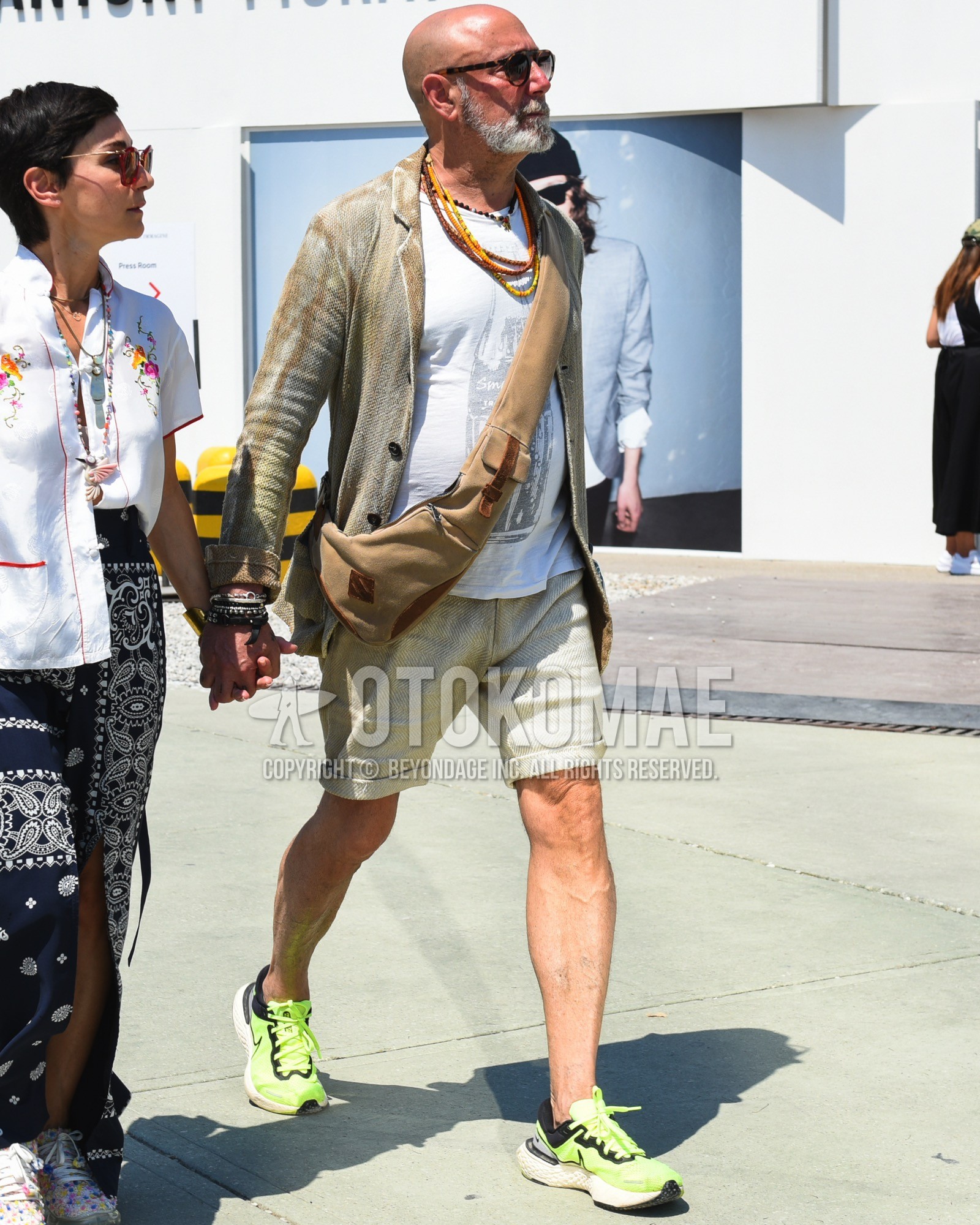 Men's spring summer outfit with brown tortoiseshell sunglasses, beige outerwear tailored jacket, white plain t-shirt, beige plain short pants, yellow low-cut sneakers, brown plain body bag.