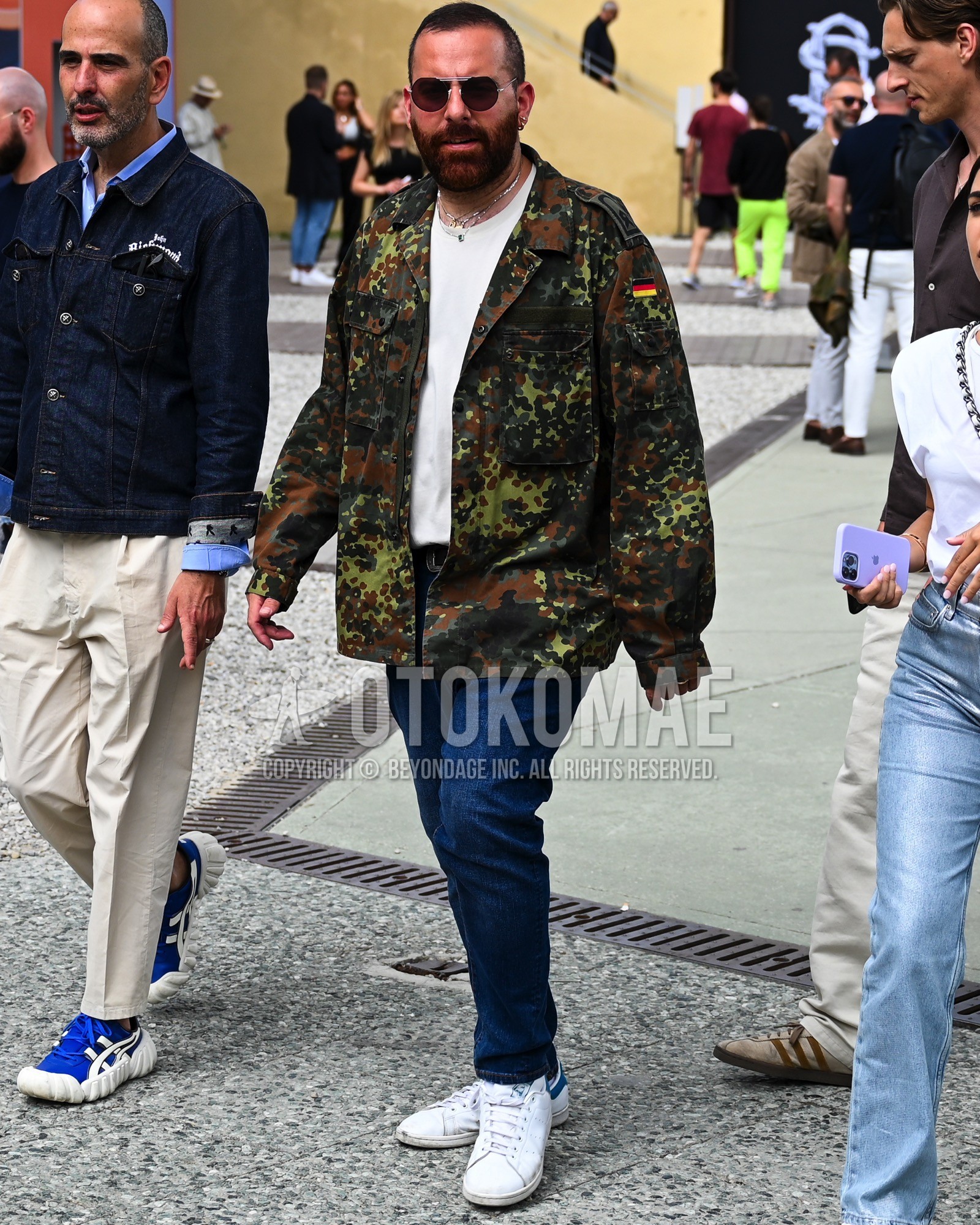 Men's spring summer autumn outfit with black plain sunglasses, olive green camouflage military jacket, white plain t-shirt, brown plain denim/jeans, white low-cut sneakers.
