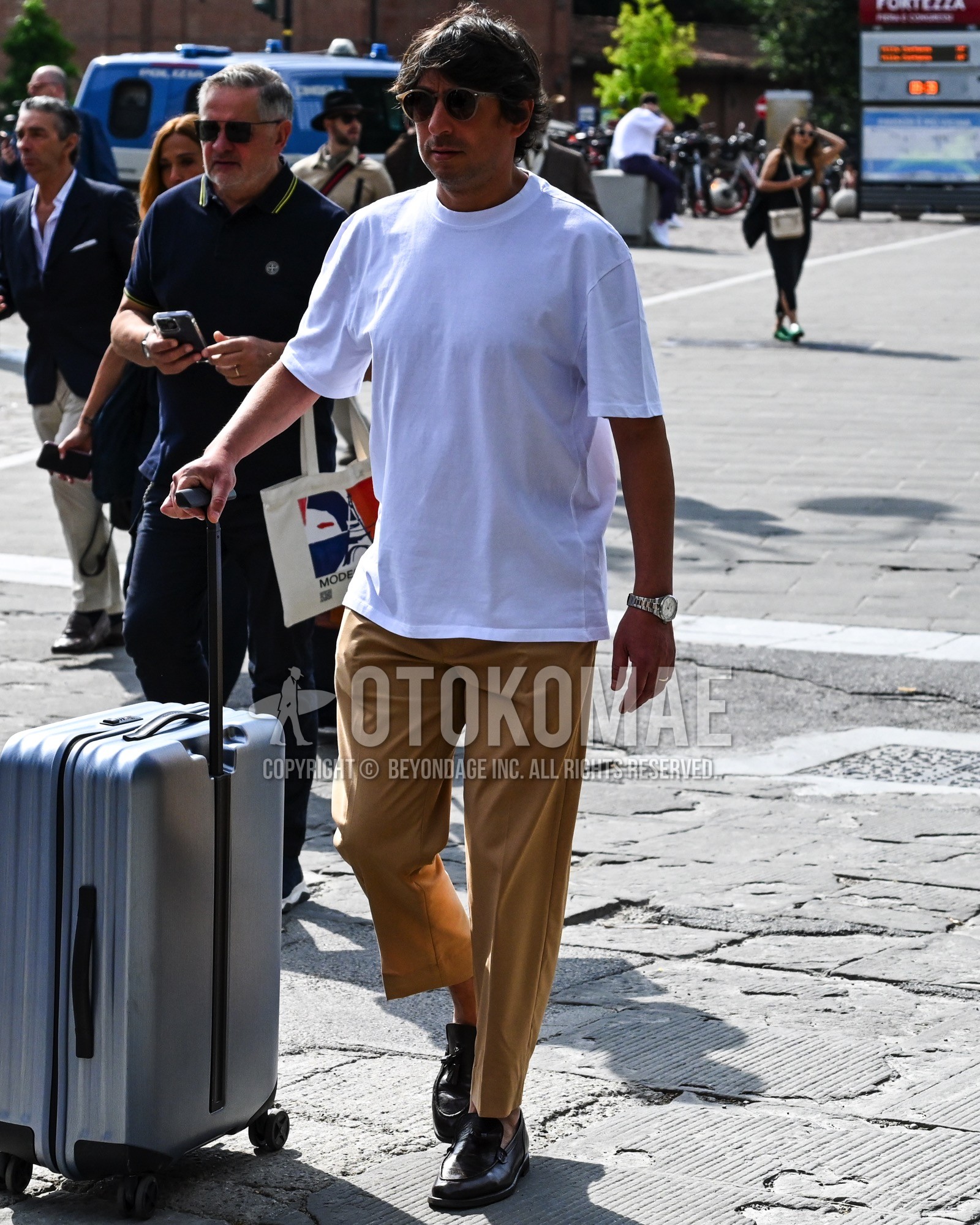 Men's spring summer outfit with black plain sunglasses, white plain t-shirt, beige plain chinos, black coin loafers leather shoes, silver plain suitcase.