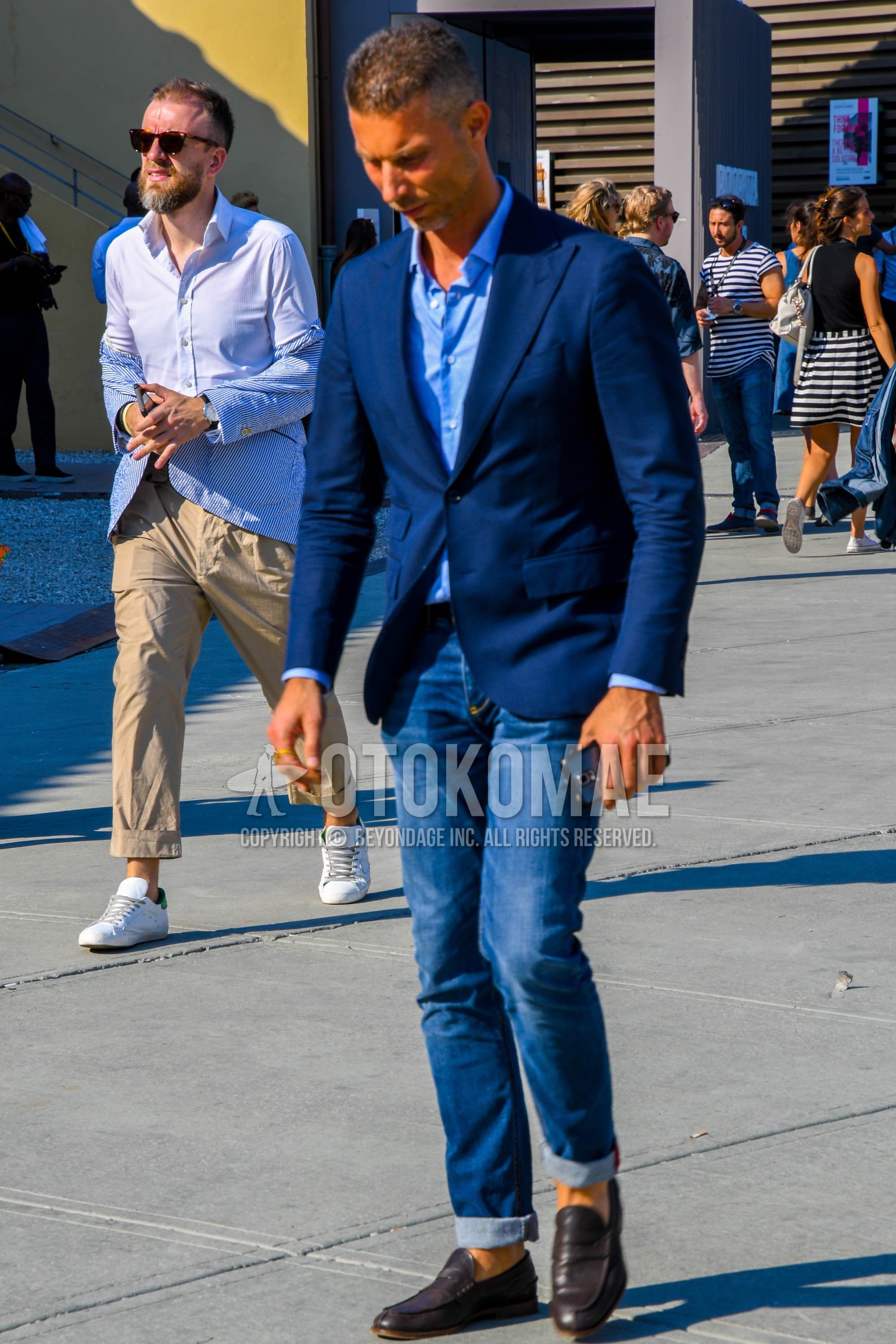 Men's spring summer autumn outfit with navy plain tailored jacket, light blue plain shirt, blue plain denim/jeans, brown coin loafers leather shoes.