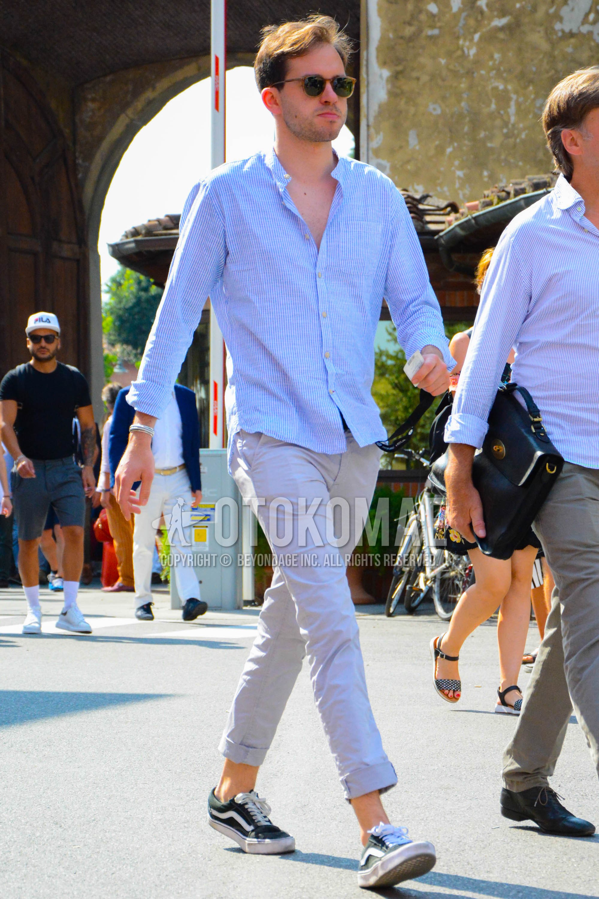 Men's spring summer outfit with brown plain sunglasses, blue stripes shirt, beige plain chinos, black low-cut sneakers.