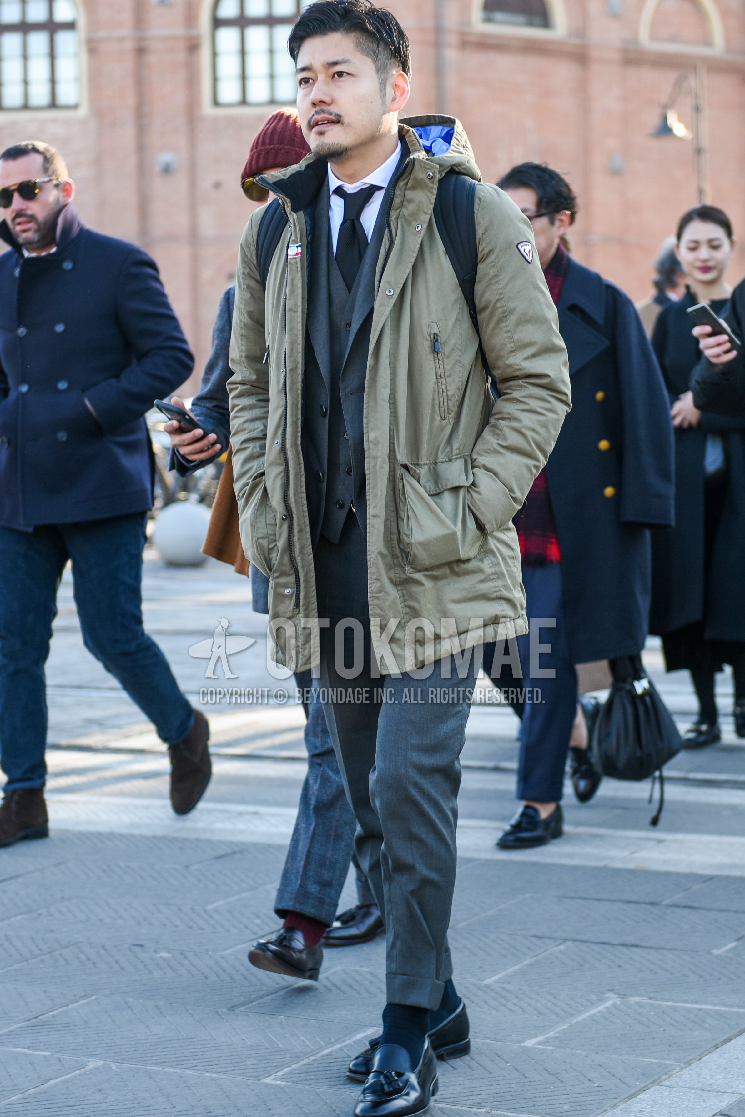 Men's winter outfit with beige plain hooded coat, white plain shirt, black plain socks, black tassel loafers leather shoes, gray plain three-piece suit, black plain necktie.