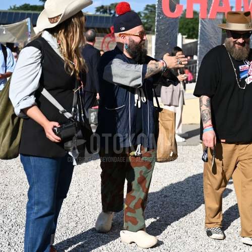 ネイビー無地のニットキャップ、オレンジべっ甲のサングラス、ネイビー無地のデニム/シャンブレーシャツ、ネイビー無地のTシャツ、オリーブグリーン・ブラウン迷彩のイージーパンツ、オリーブグリーン・ブラウン迷彩のスキニーパンツ、白ビーチサンダル、ベージュ無地のトートバッグを合わせた春夏のメンズコーデ・着こなし。