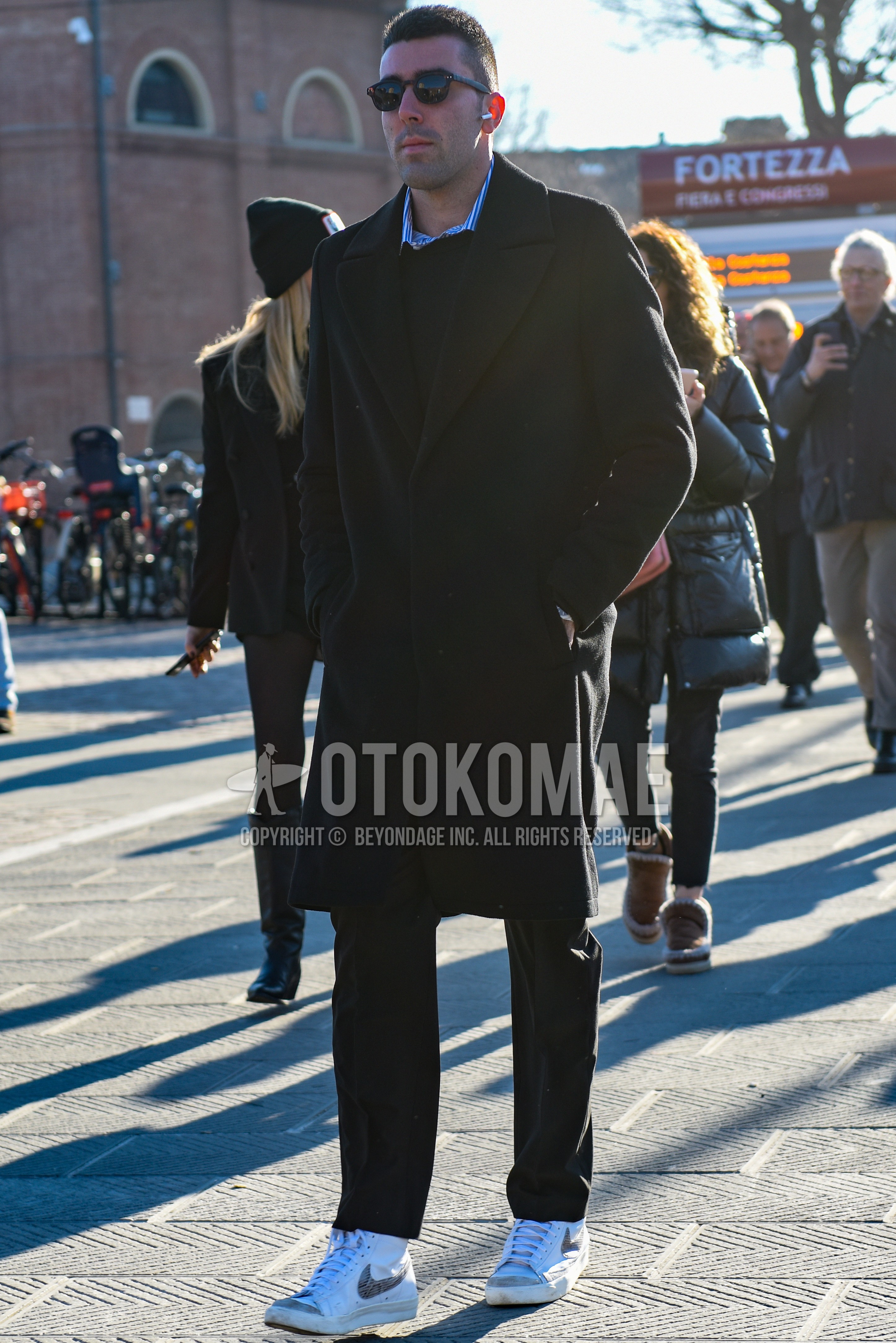 Men's autumn winter outfit with brown tortoiseshell sunglasses, black plain chester coat, black plain sweater, blue stripes shirt, dark gray plain slacks, white high-cut sneakers.