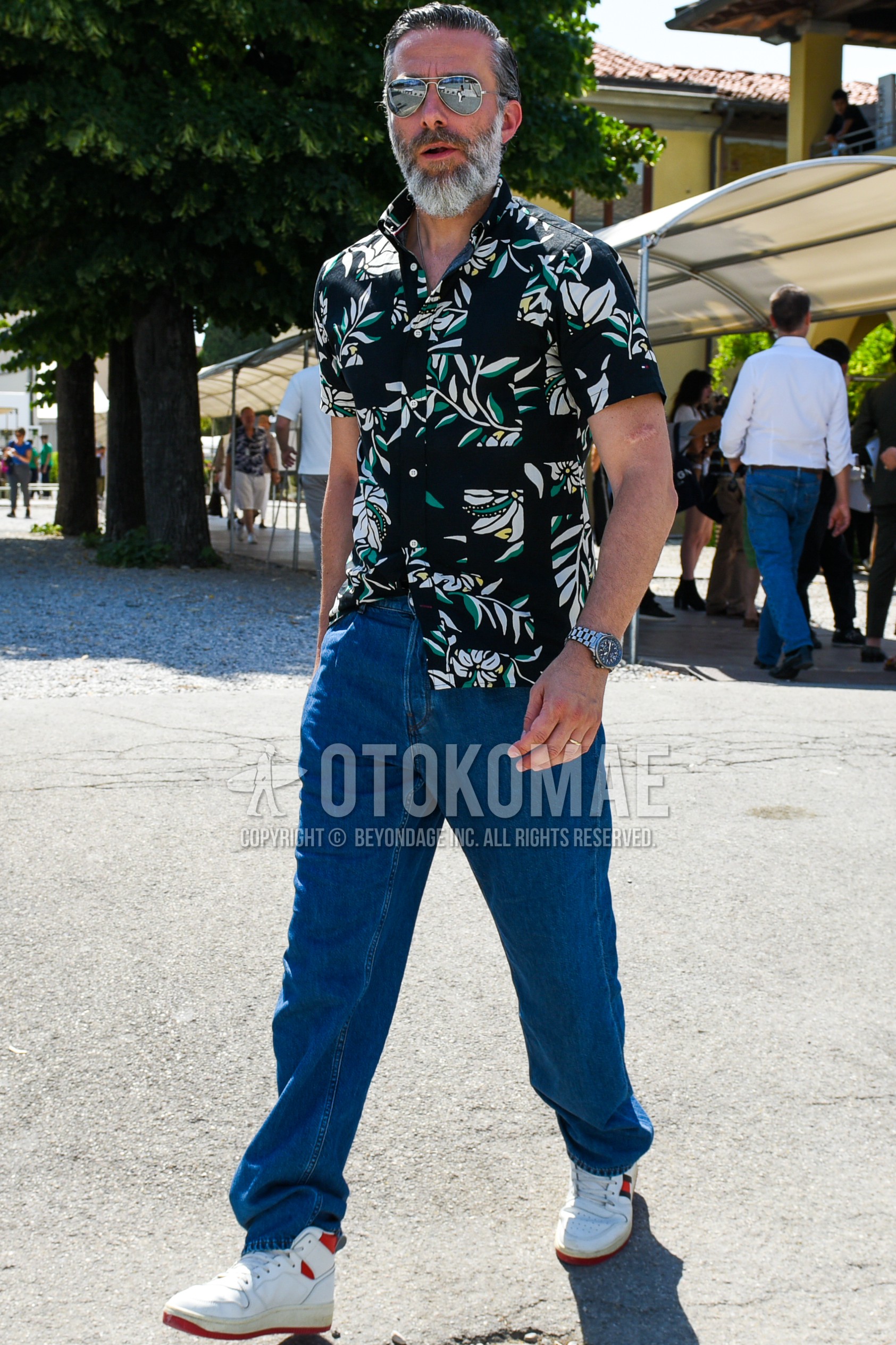 Men's spring summer outfit with silver plain sunglasses, black botanical shirt, blue plain denim/jeans, white high-cut sneakers.