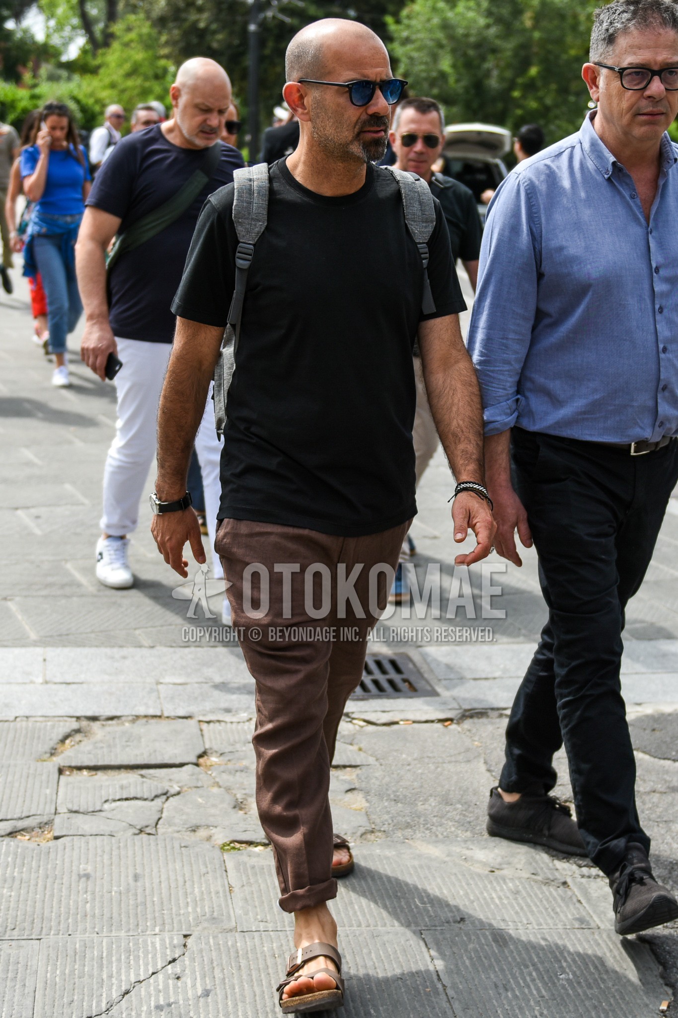 Men's summer outfit with black plain sunglasses, black plain t-shirt, brown plain slacks, brown leather sandals.