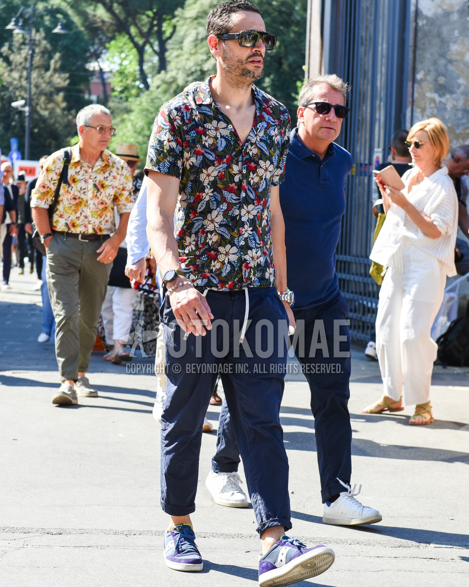 Men's spring summer outfit with olive green tortoiseshell sunglasses, olive green botanical shirt, navy plain easy pants, navy plain cropped pants, purple low-cut sneakers.