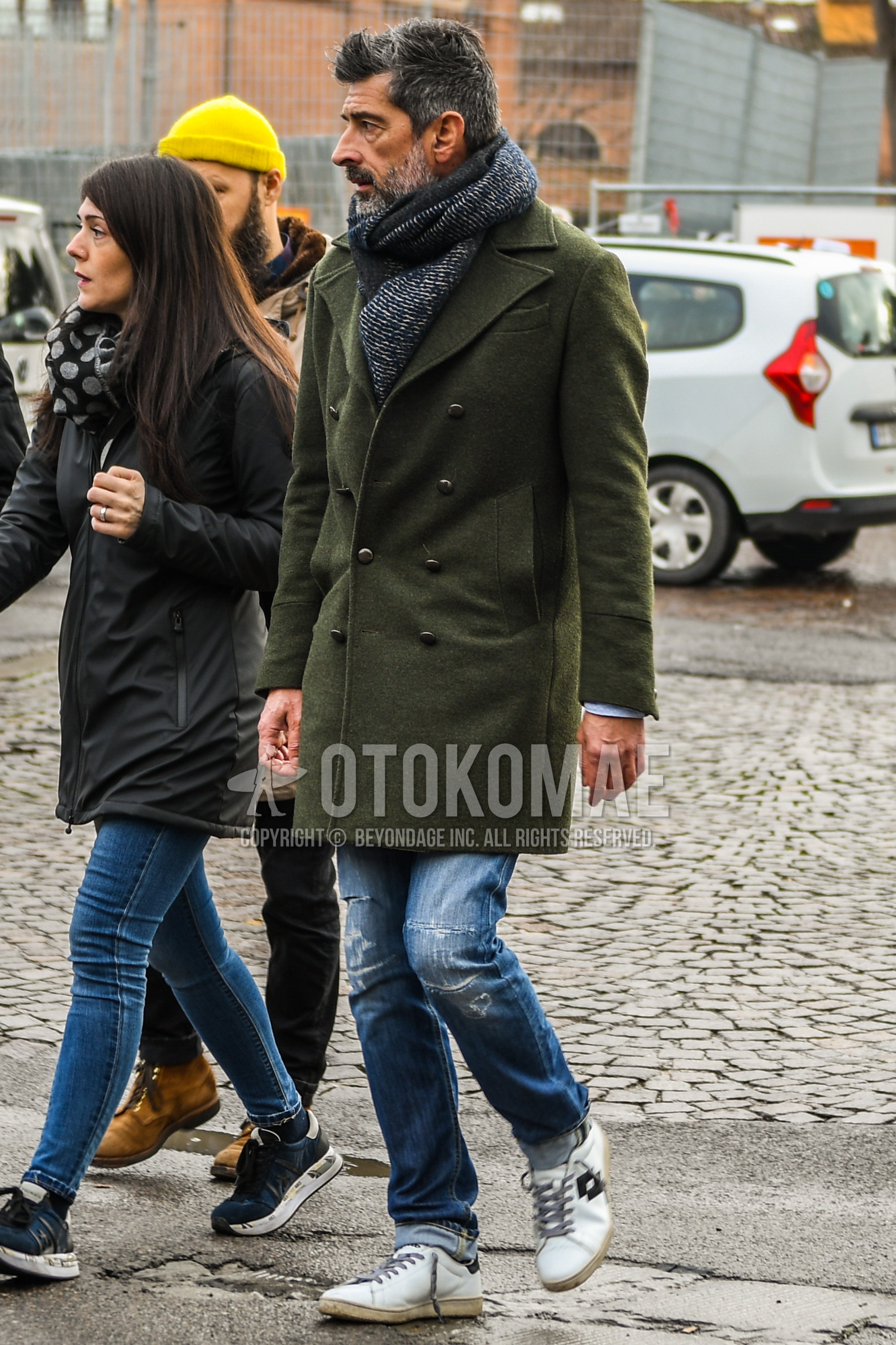 Men's winter outfit with dark gray plain scarf, olive green plain chester coat, blue plain damaged jeans, white low-cut sneakers.