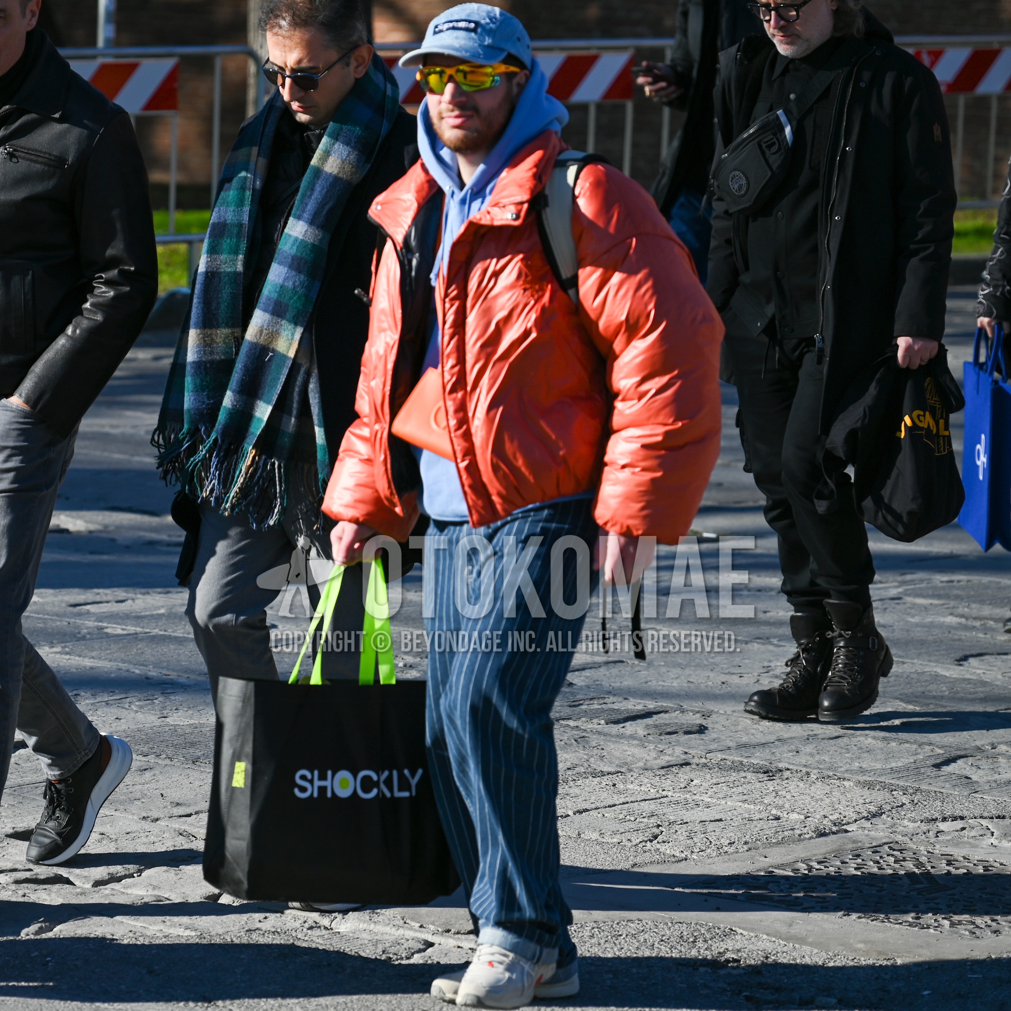 Men's winter outfit with light blue plain baseball cap, multi-color eyewear sunglasses, orange plain down jacket, purple plain hoodie, navy stripes slacks, gray low-cut sneakers, black plain bag.