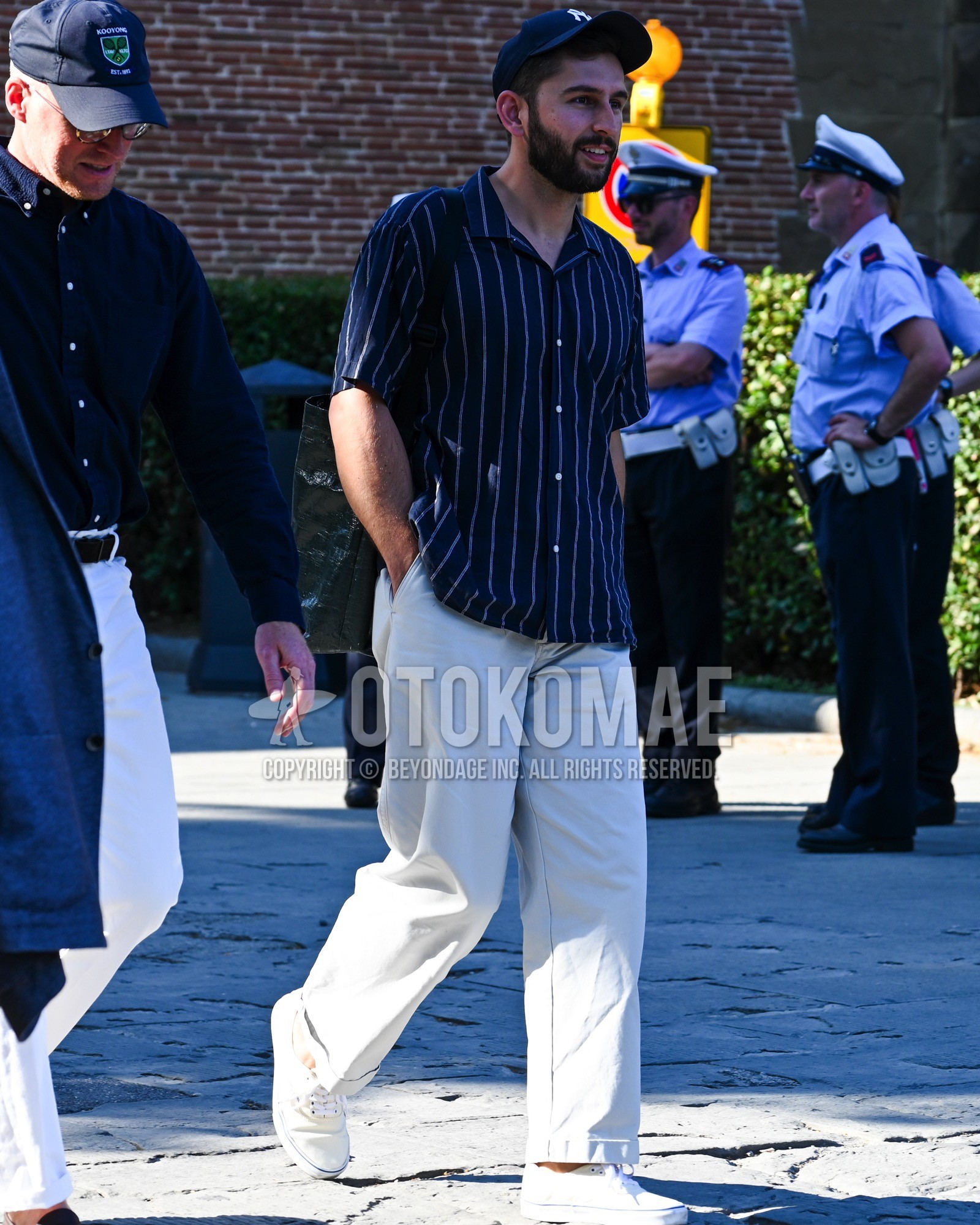 Men's spring summer outfit with navy deca logo baseball cap, navy stripes shirt, white plain chinos, white low-cut sneakers.
