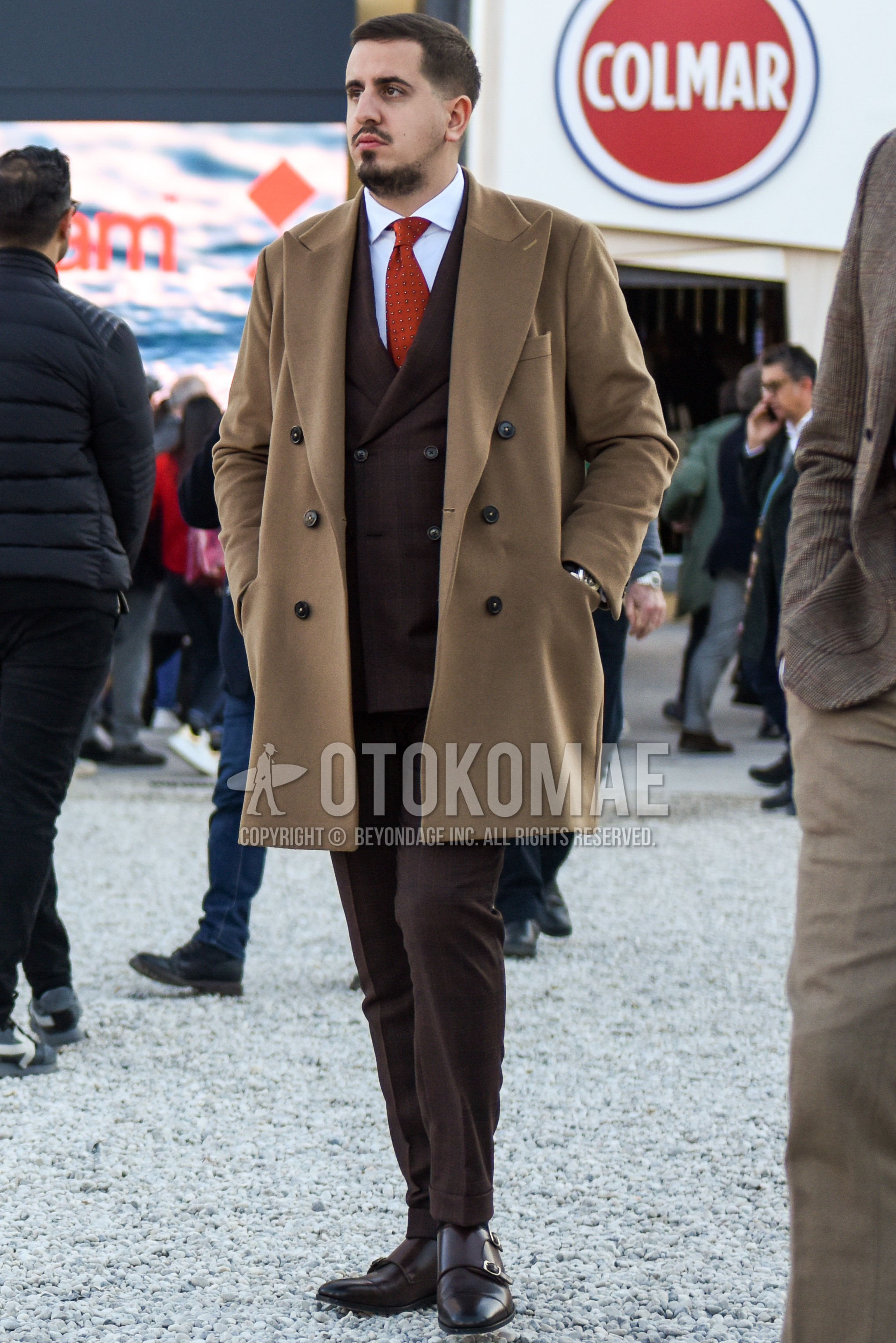 Men's autumn winter outfit with beige plain chester coat, white plain shirt, brown monk shoes leather shoes, brown check suit, orange necktie necktie.