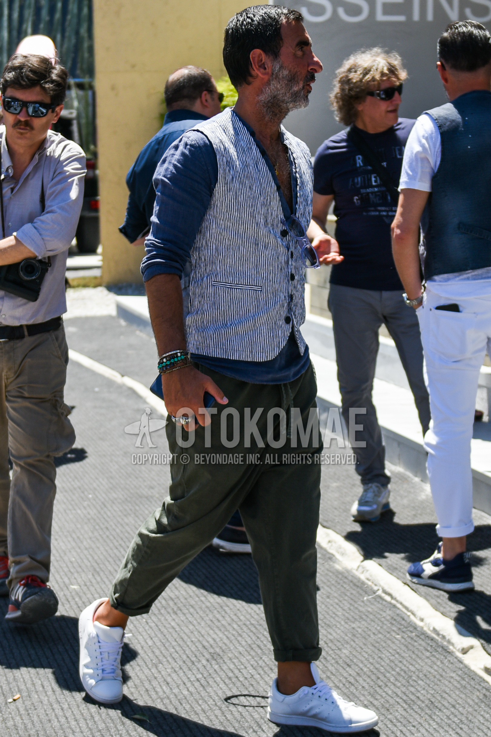 Men's spring summer outfit with white blue stripes gilet, gray plain shirt, olive green plain easy pants, plain chinos, white low-cut sneakers.