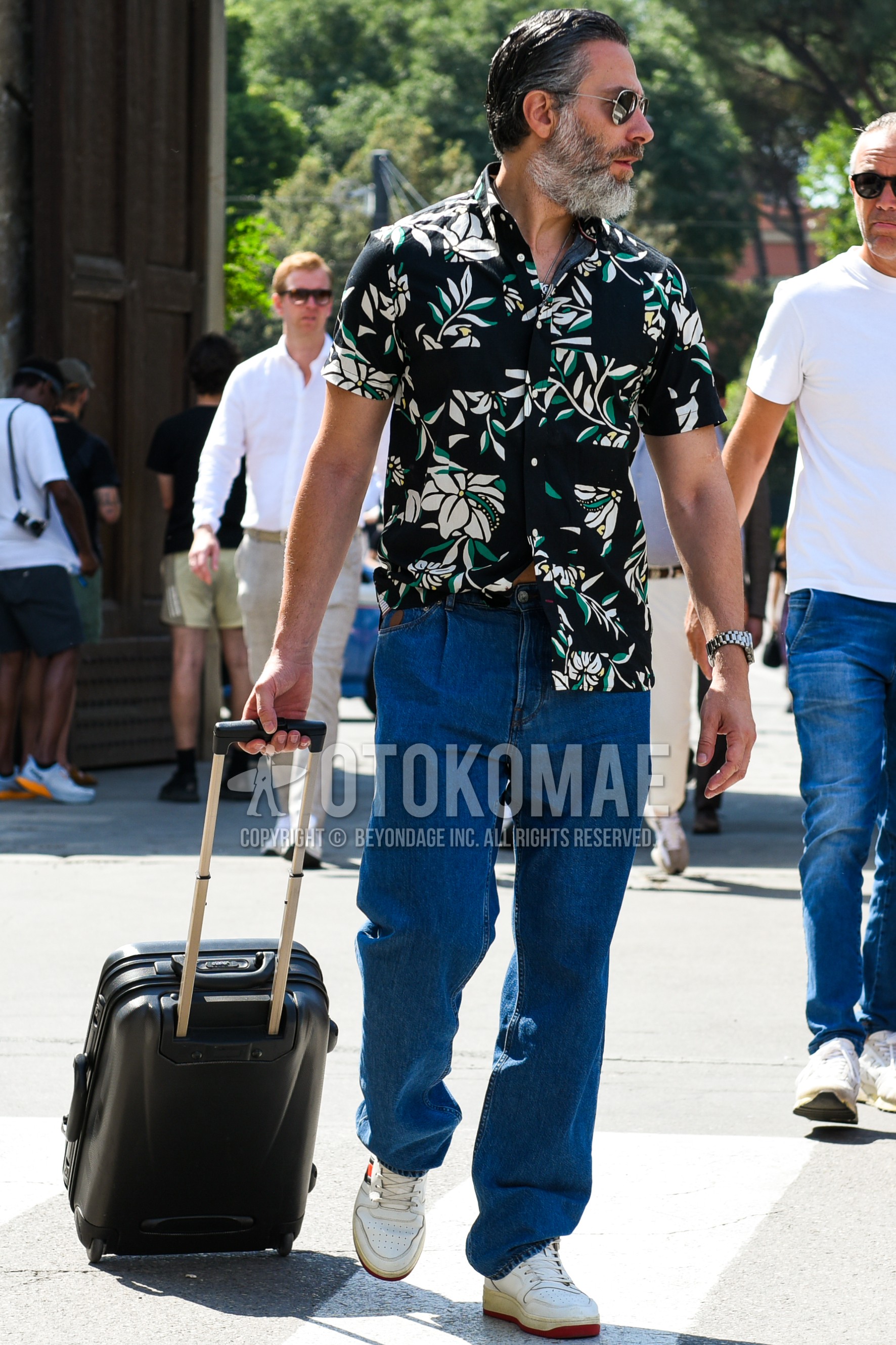 Men's spring summer outfit with silver plain sunglasses, black botanical shirt, blue plain denim/jeans, white low-cut sneakers, black plain bag.