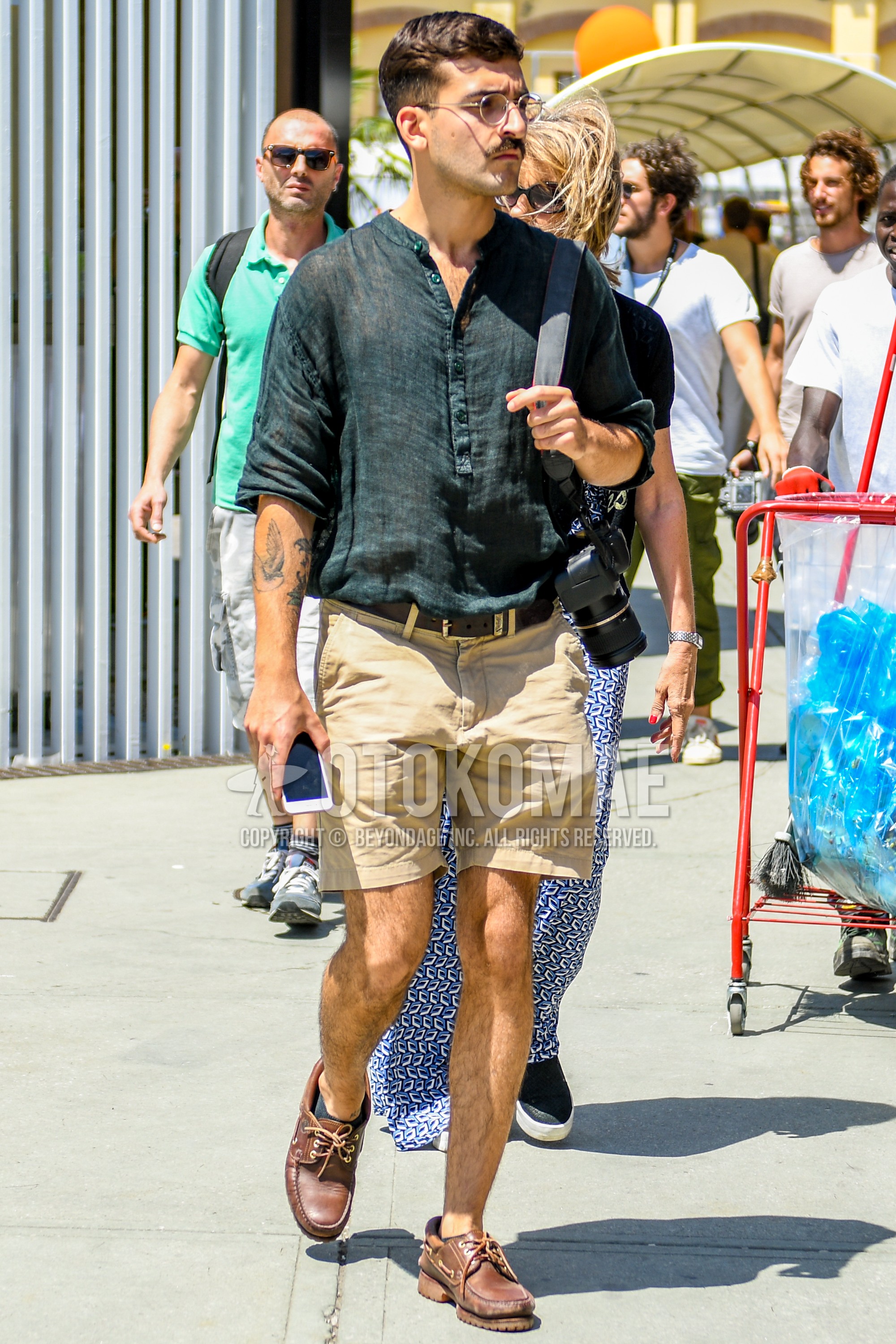 Men's summer outfit with plain glasses, black plain shirt, brown plain leather belt, beige plain short pants, plain chinos, brown moccasins/deck shoes leather shoes.