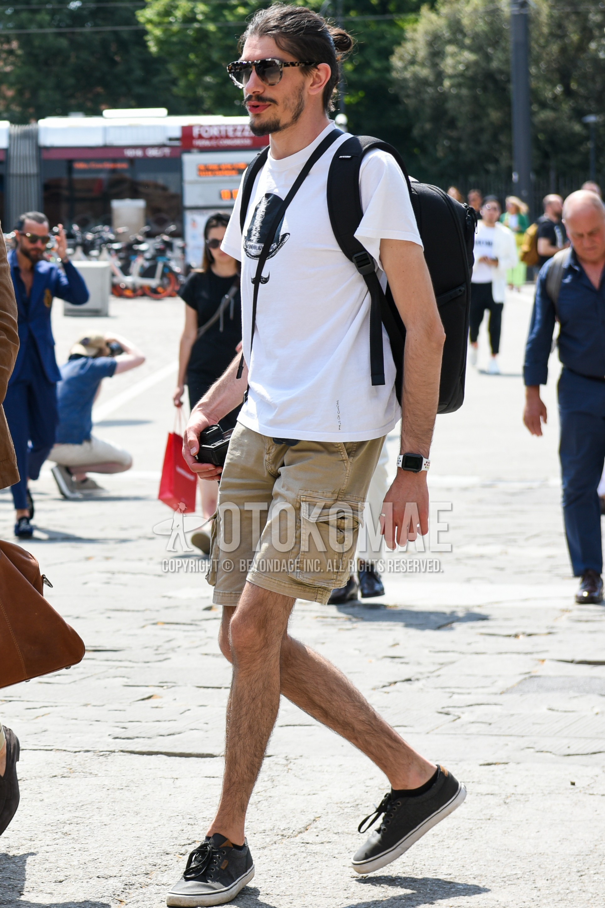 Men's summer outfit with silver tortoiseshell sunglasses, white graphic t-shirt, beige plain short pants, beige plain cargo pants, dark gray low-cut sneakers.